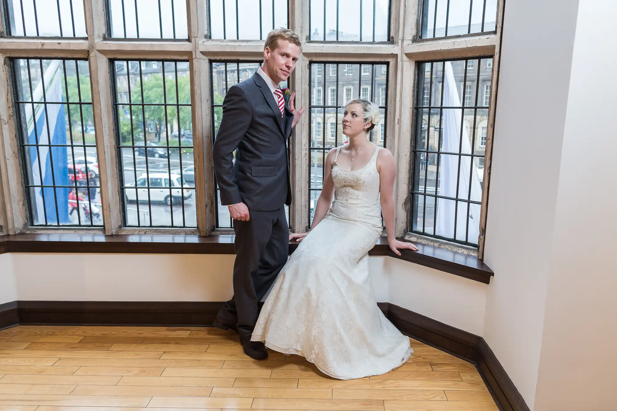 A bride in a white gown and a groom in a suit pose by large windows in a room with wooden floors.