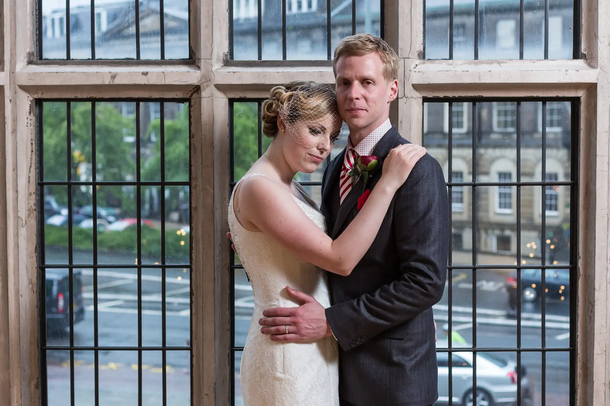 A newlywed couple embracing by a large window, with the woman in a white dress leaning against the man in a suit, overlooking a city street.