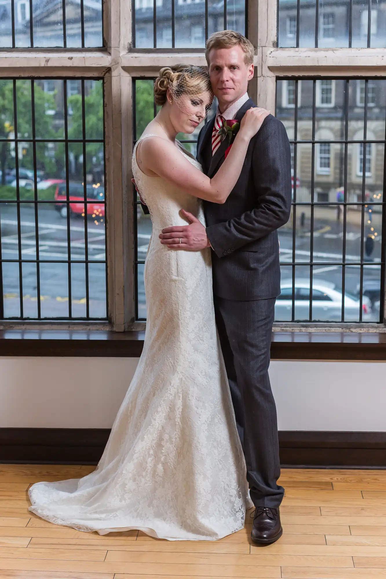 A newlywed couple posing affectionately in front of a large window with street views, the bride in an elegant lace gown and the groom in a black suit with a red boutonniere.