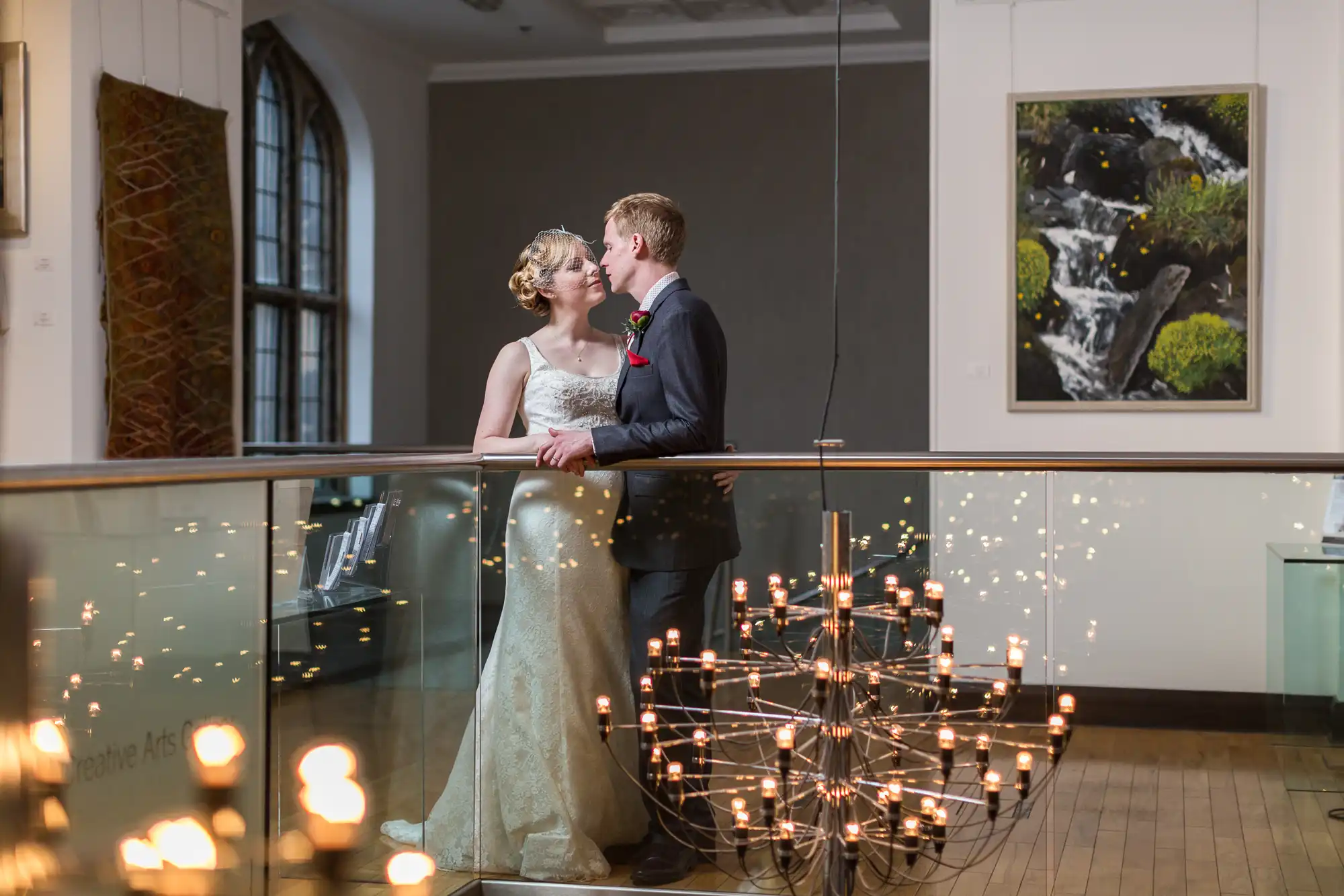 A newlywed couple shares an intimate moment in an art gallery, standing next to a sparkling chandelier with artwork in the background.
