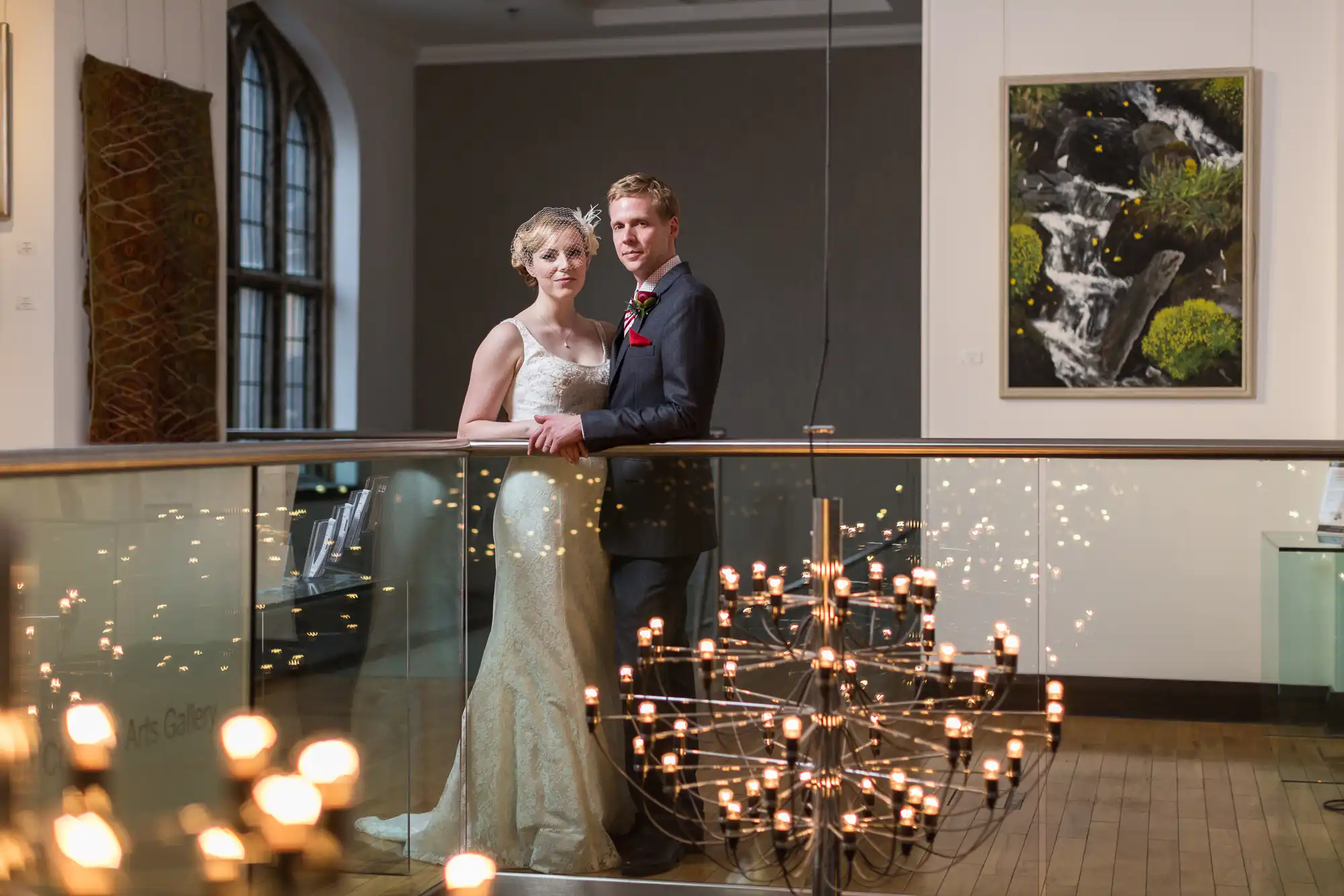 A bride and groom standing together in a museum, with a modern chandelier in the foreground and paintings in the background.