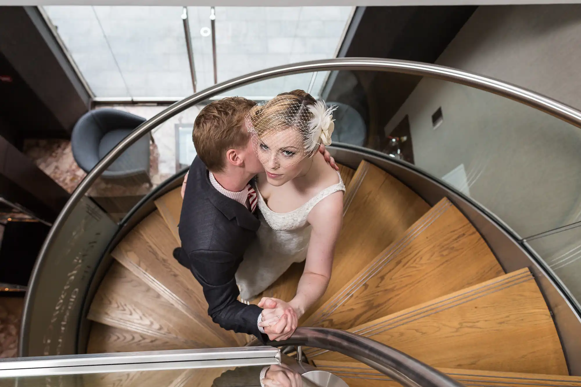 A bride and groom share a kiss on a spiral staircase, viewed from above.