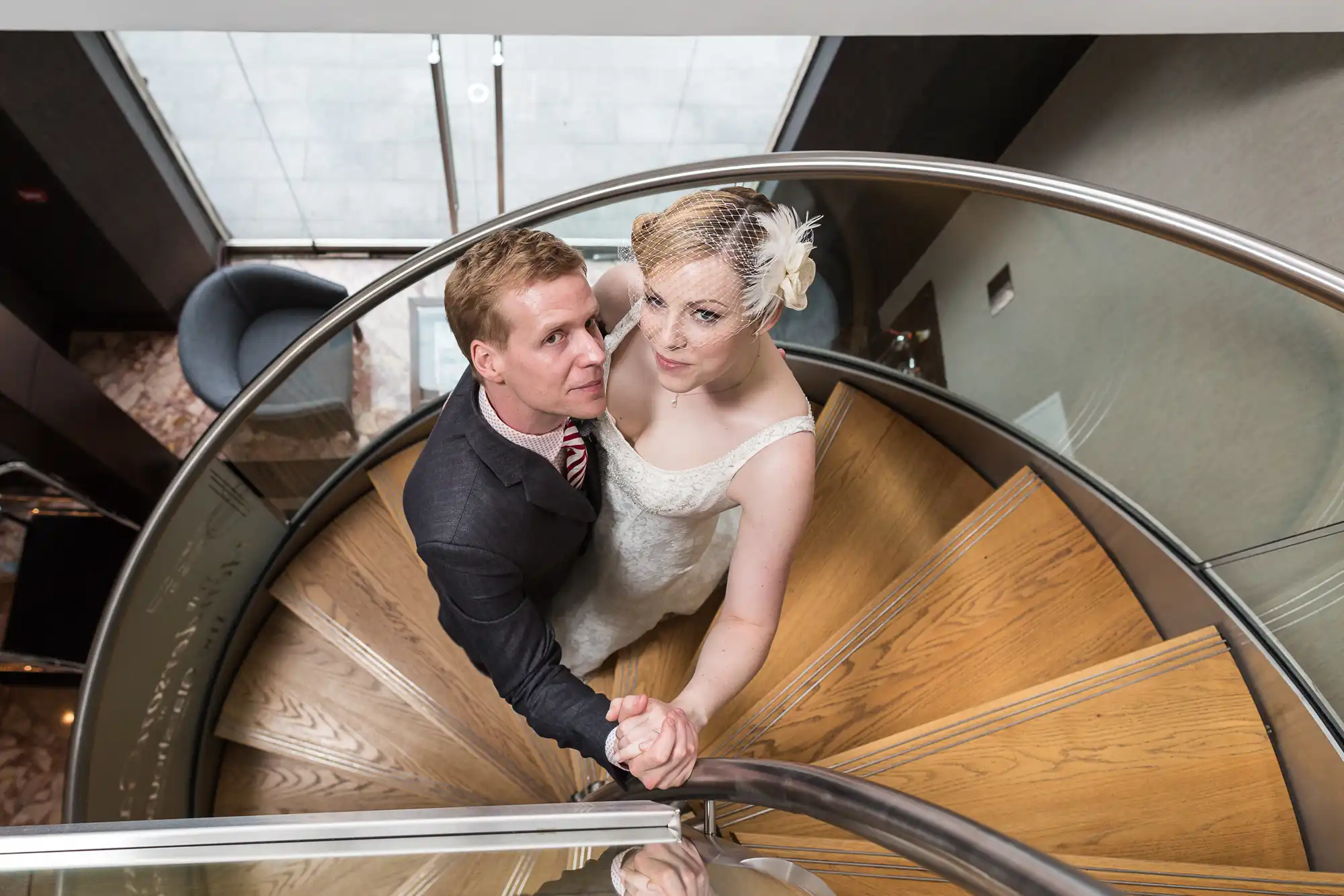 A newlywed couple holding hands while ascending a spiral staircase, looking upwards towards the camera.