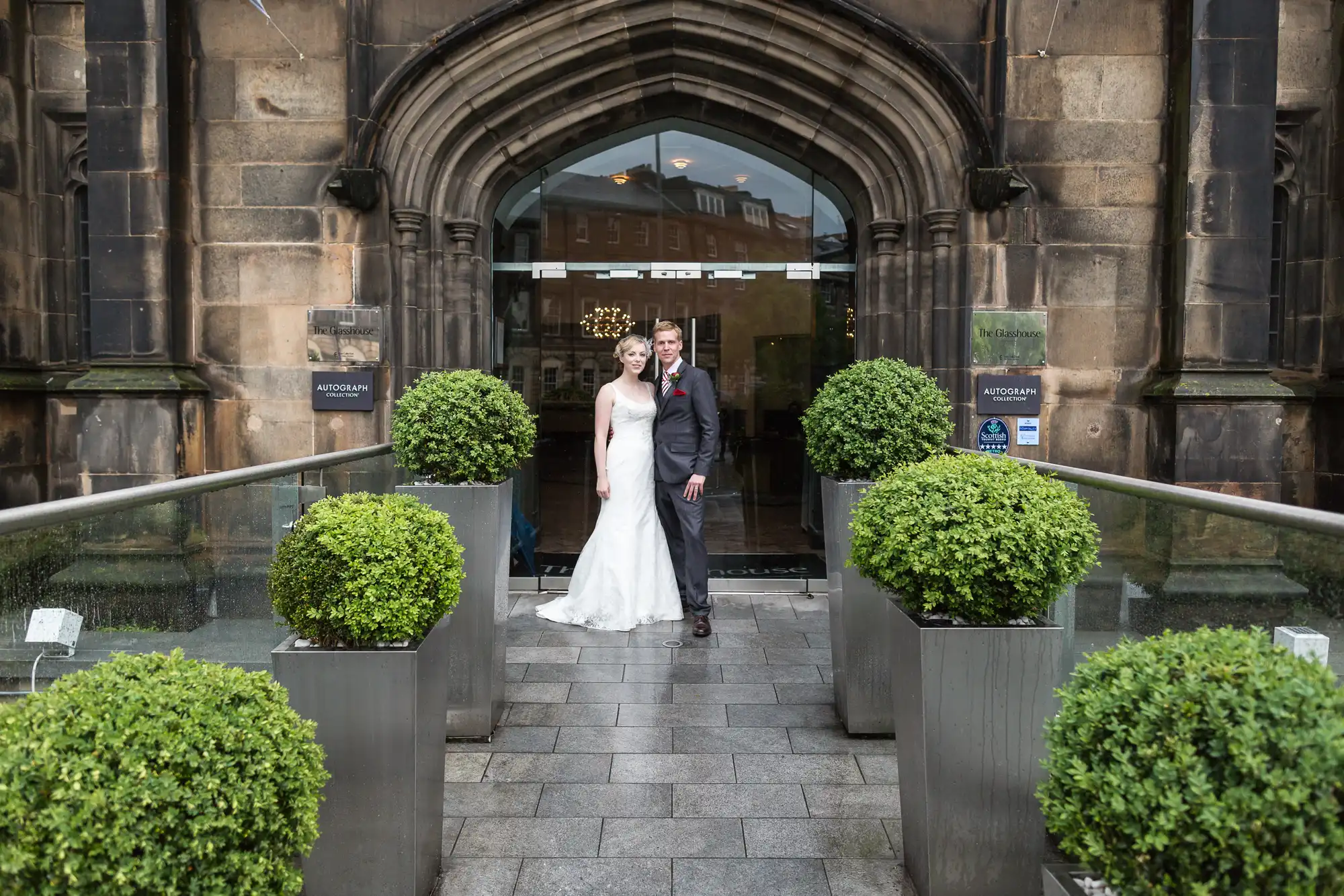 A bride and groom walking out of a church entrance, smiling and holding hands, with topiary plants lining the pathway.