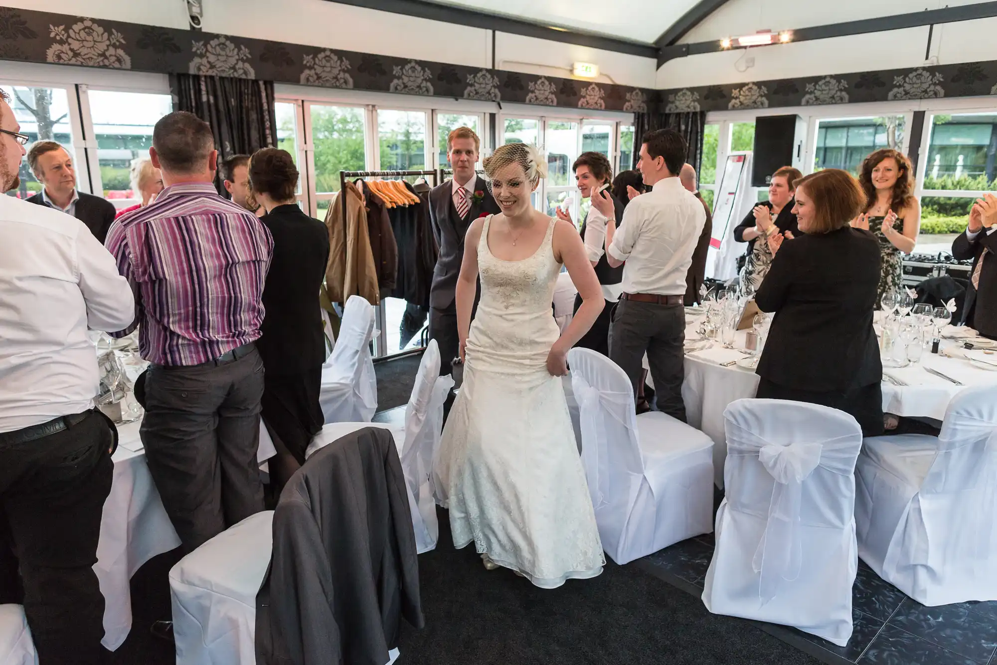 A bride in a white gown walking through a busy wedding reception hall filled with guests and adorned tables.