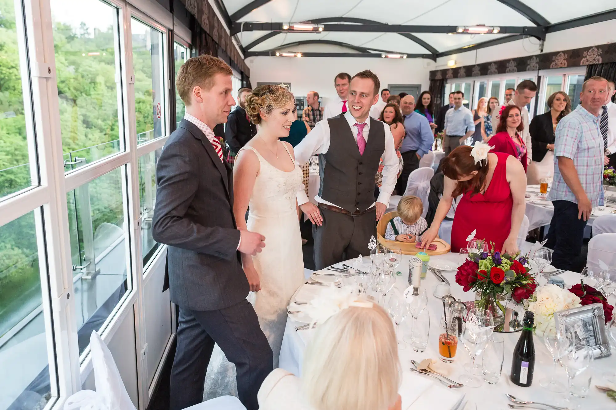 A bride and groom walking through a crowded reception hall with guests around tables adorned with red floral arrangements.