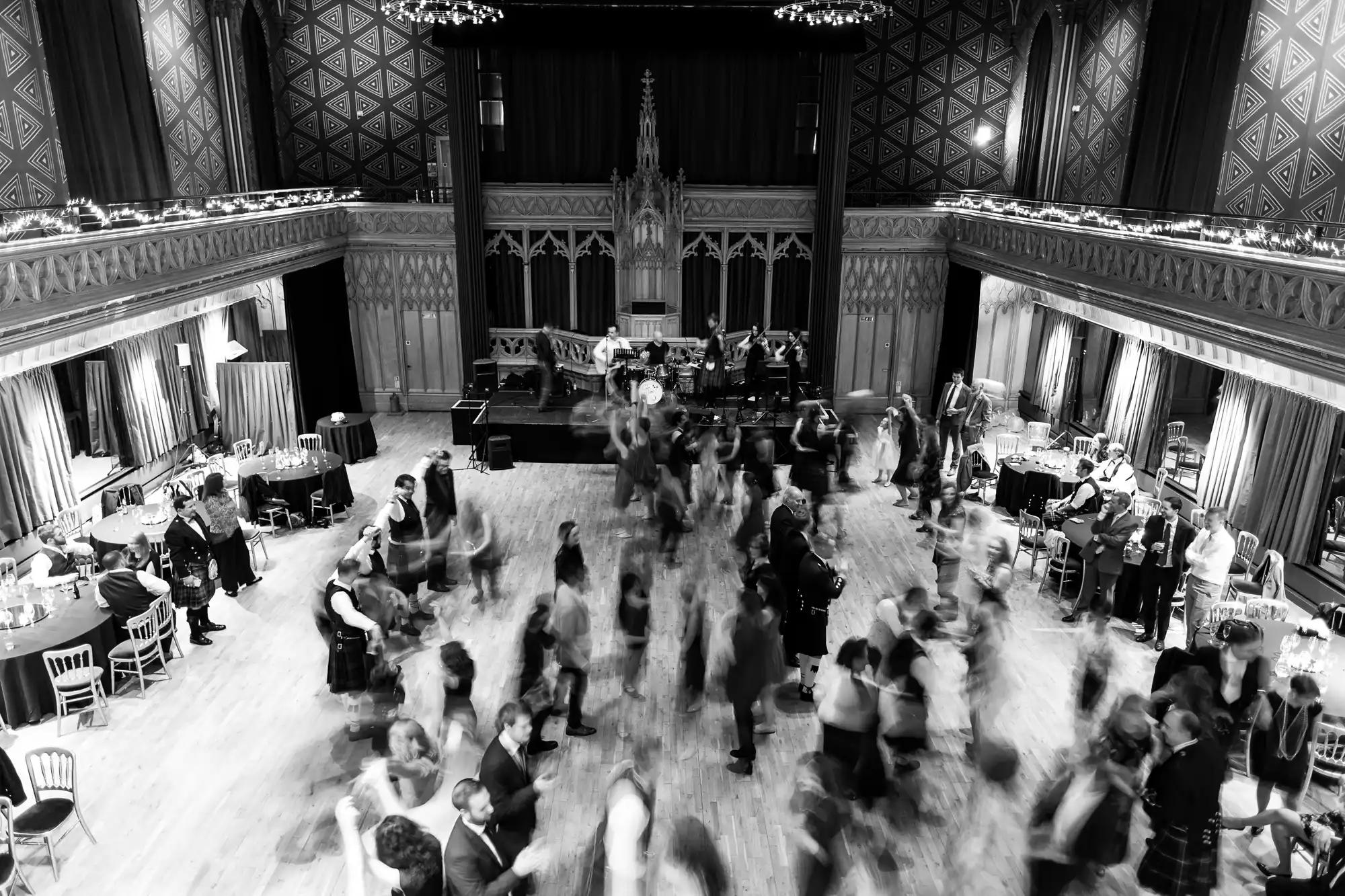 Black and white photo of a ballroom with many people dancing on the floor and musicians on stage. The audience is seated around the room. The image captures motion blur from dancers.