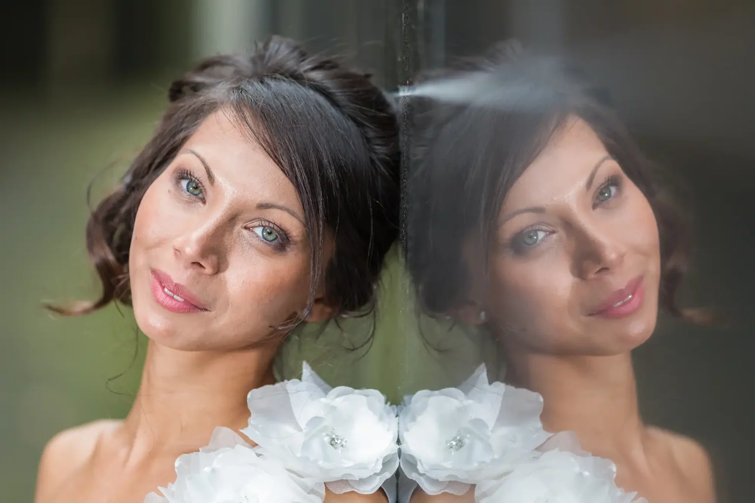 A woman with brown hair and white floral accessories poses beside a reflective glass surface, showing her reflection.