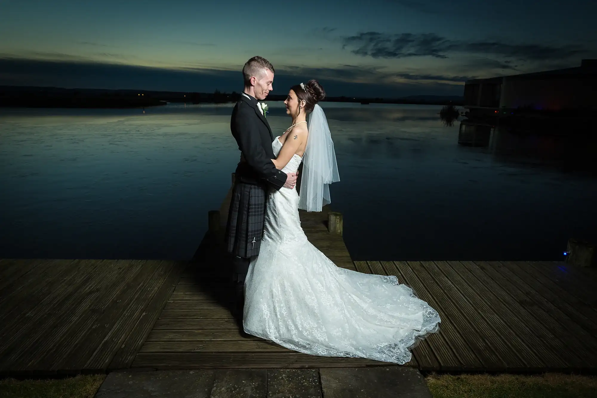 A bride and groom embrace on a lakeside dock at dusk, their figures illuminated against a twilight sky.