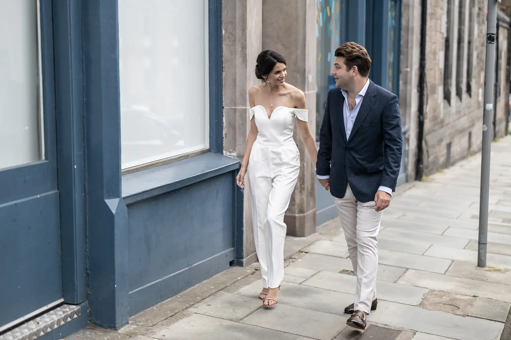 A young couple, a woman in a white jumpsuit and a man in a blue blazer and white pants, walking and smiling at each other in a city street.