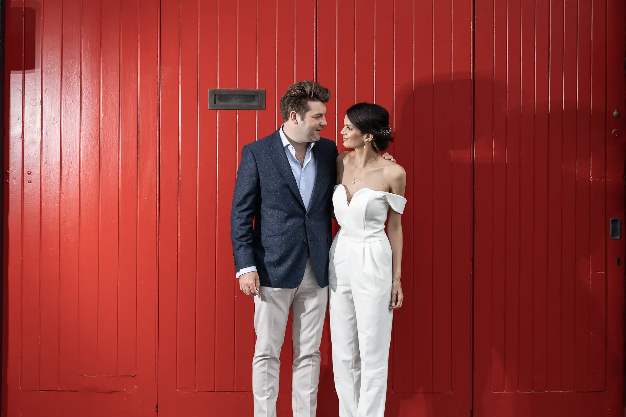 A couple, the woman in a white jumpsuit and the man in a blazer and white pants, leaning affectionately towards each other against a vibrant red wall.