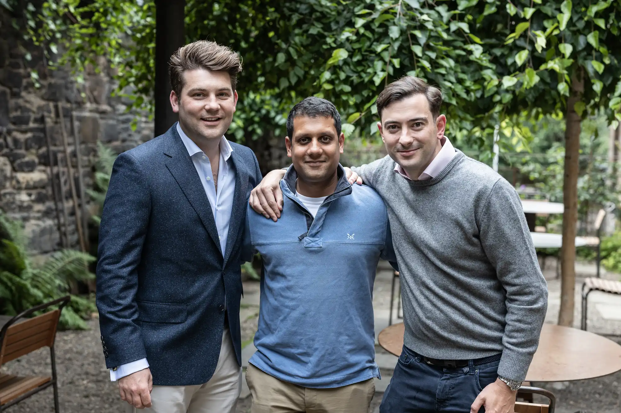 Three men smiling at the camera in a garden setting, two in sweaters and one in a polo shirt.