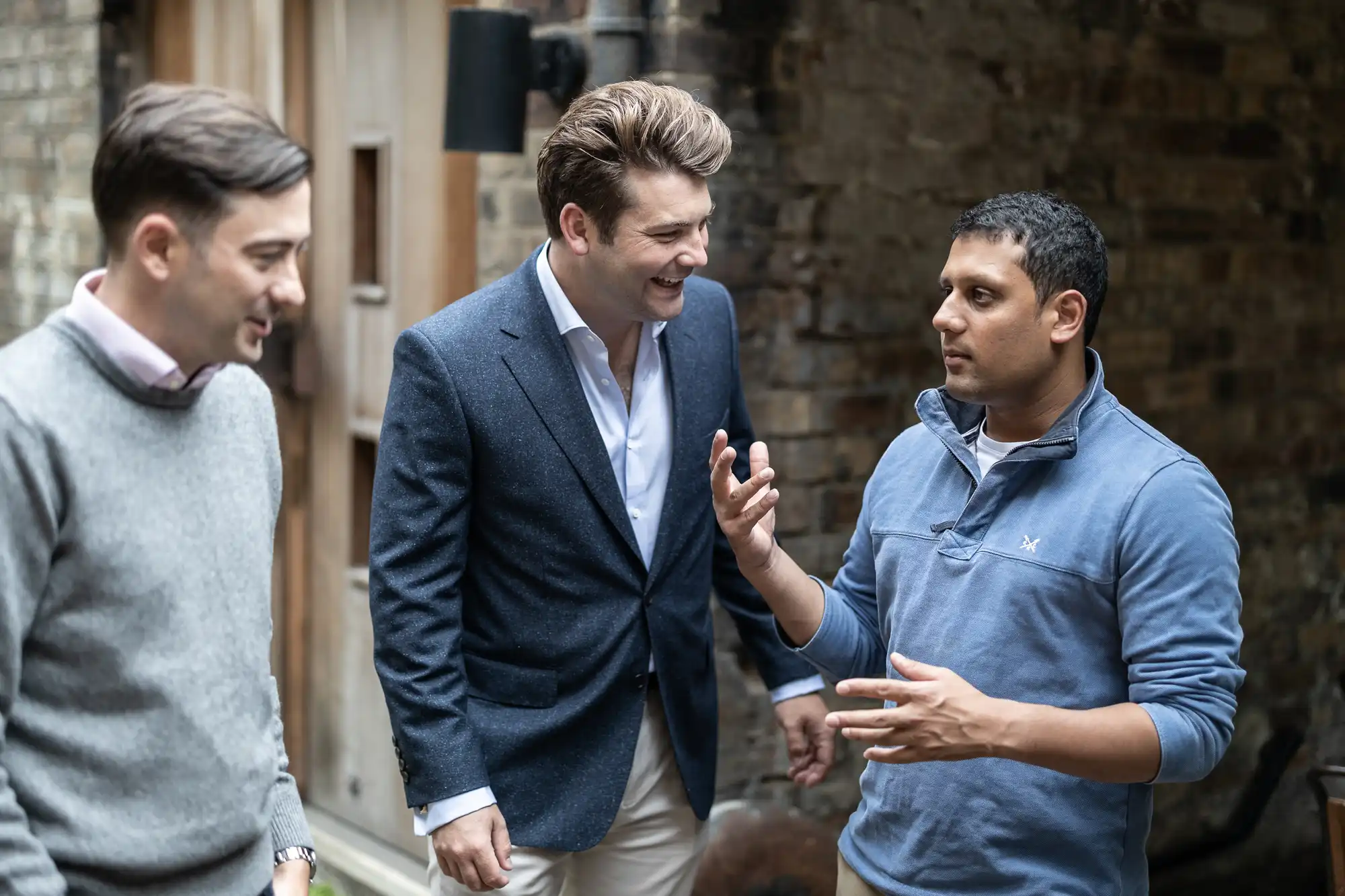 Three men engaged in a casual conversation outdoors, expressing positive emotions and gestures. two are listening intently while one speaks animatedly.