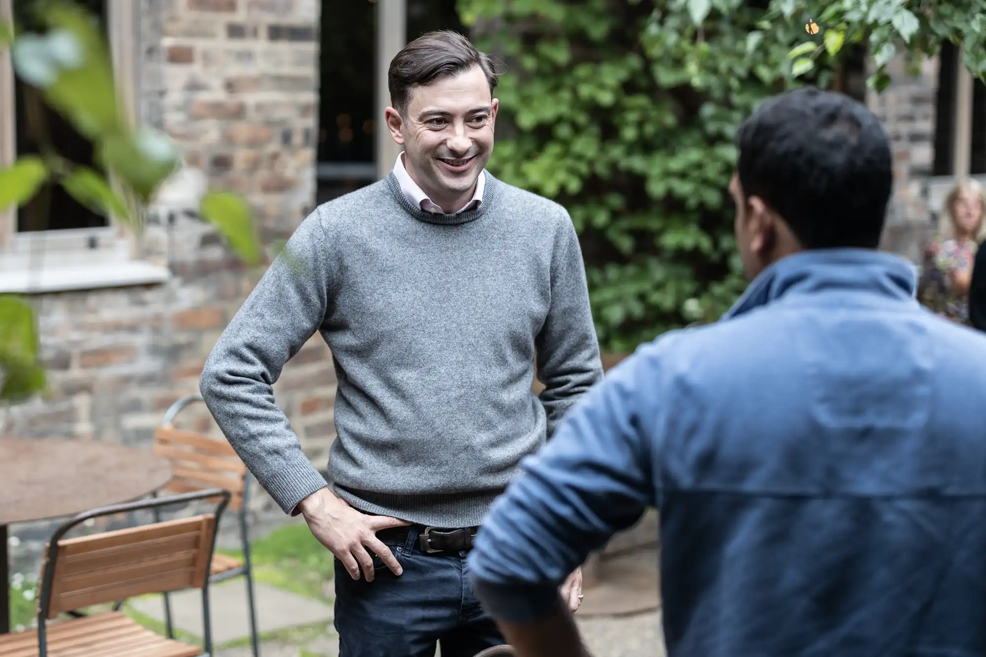 Two men standing and conversing in a courtyard, with one facing the camera and smiling, wearing a grey sweater and jeans.