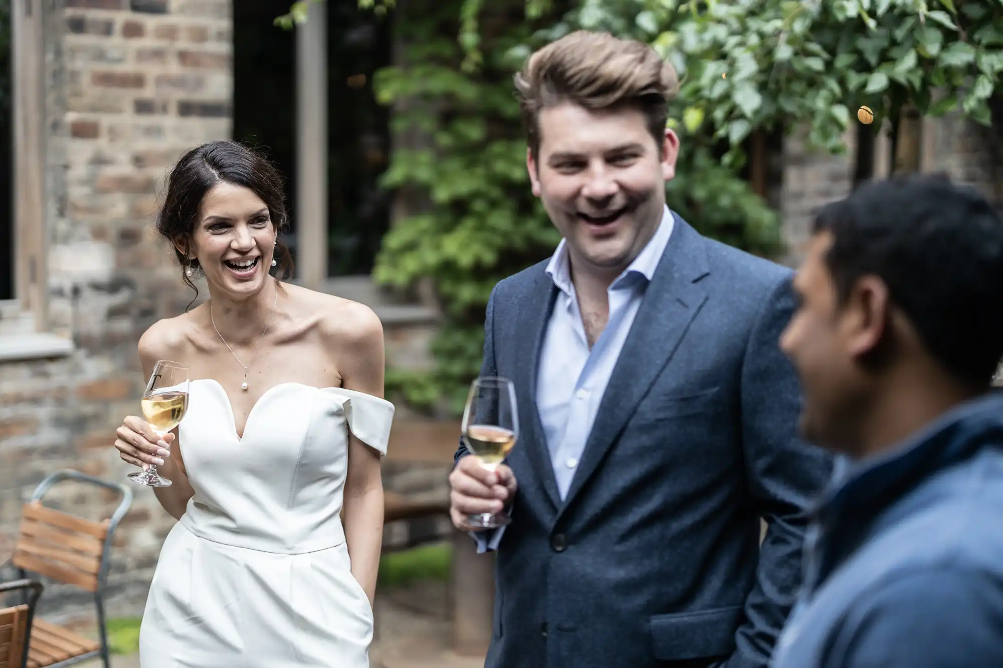 A woman in a white dress and a man in a suit laugh while holding champagne glasses, conversing with a man in a blue shirt at an outdoor gathering.