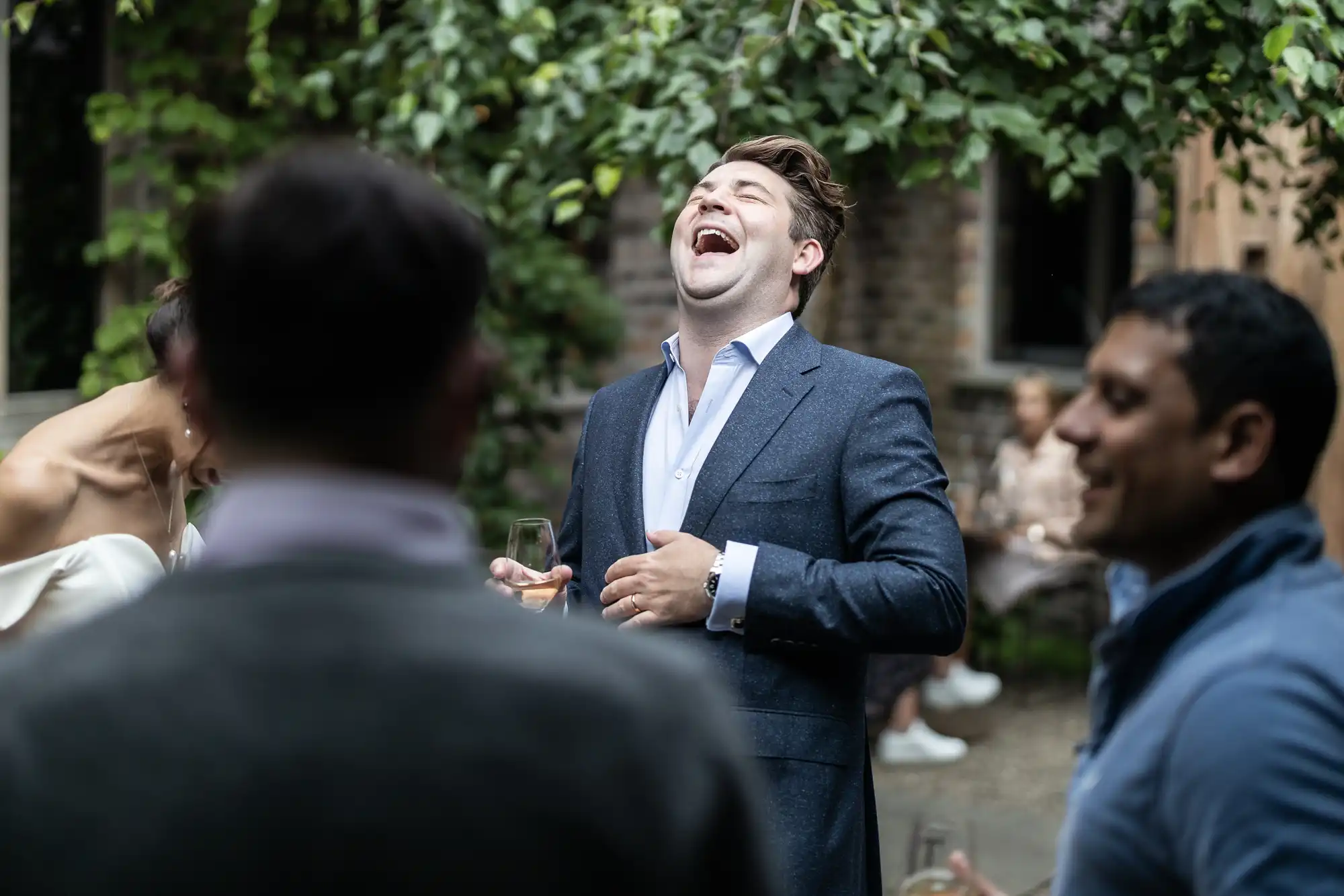 A man in a suit laughs joyfully while holding a wine glass at an outdoor gathering with other guests around him.