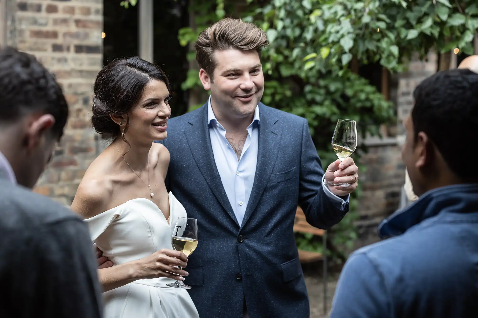 Bride and groom holding champagne glasses at a wedding reception, interacting happily with guests in an outdoor setting.