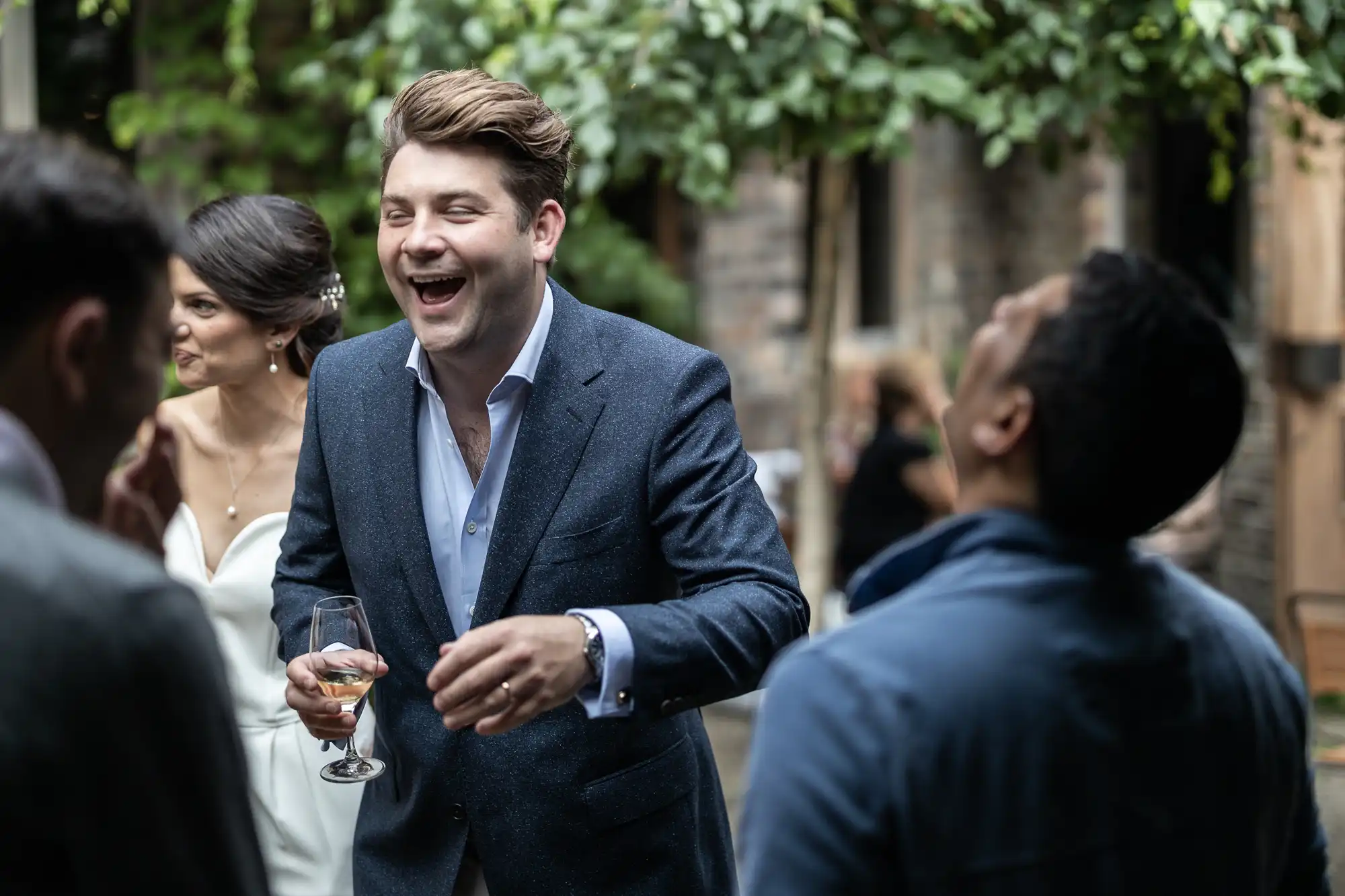 A man in a suit laughs joyfully while holding a wine glass at an outdoor wedding, with guests around him.