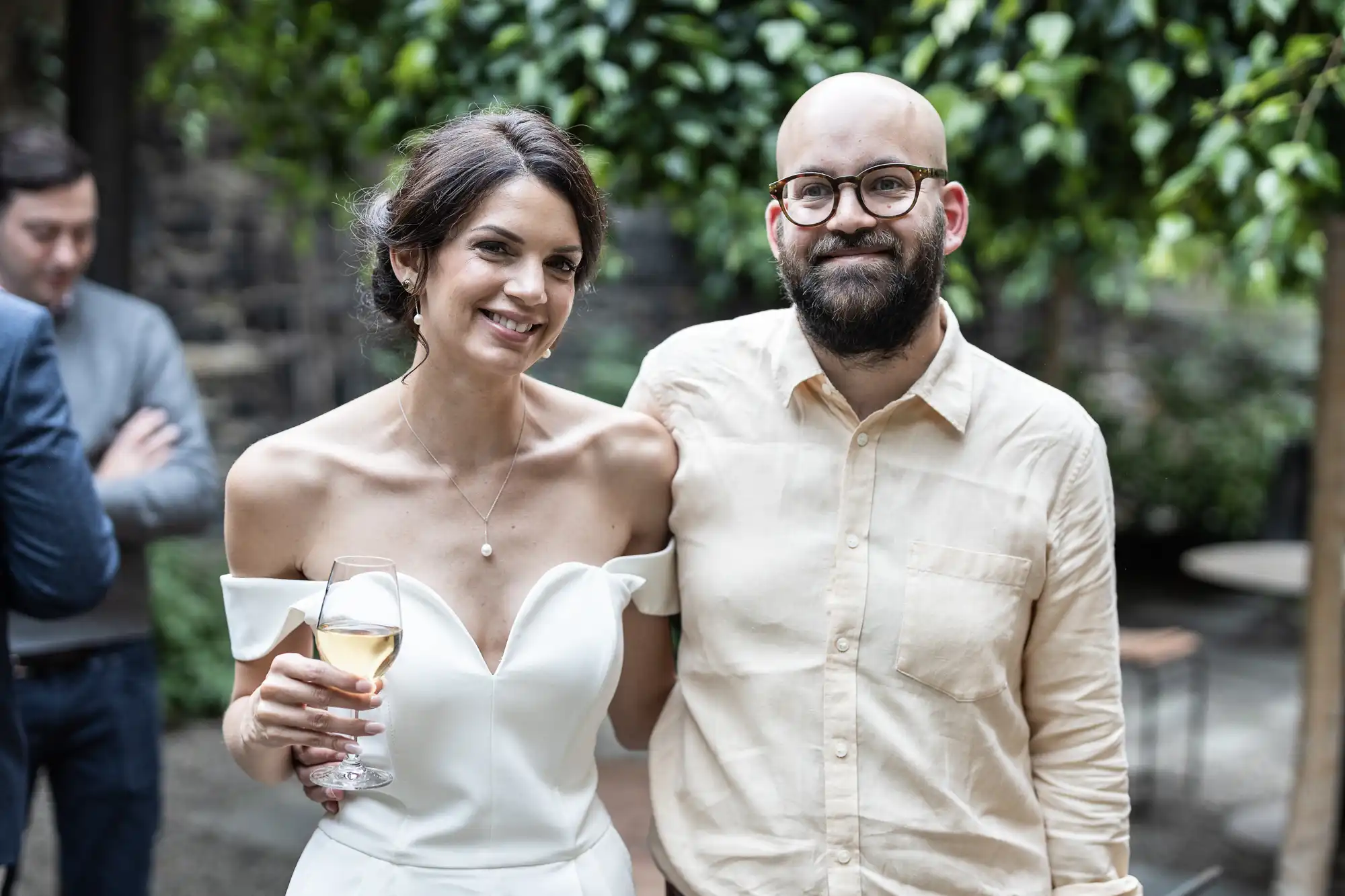 A bride in a white dress and a groom in a beige shirt, both smiling and standing outdoors with the bride holding a glass of wine.