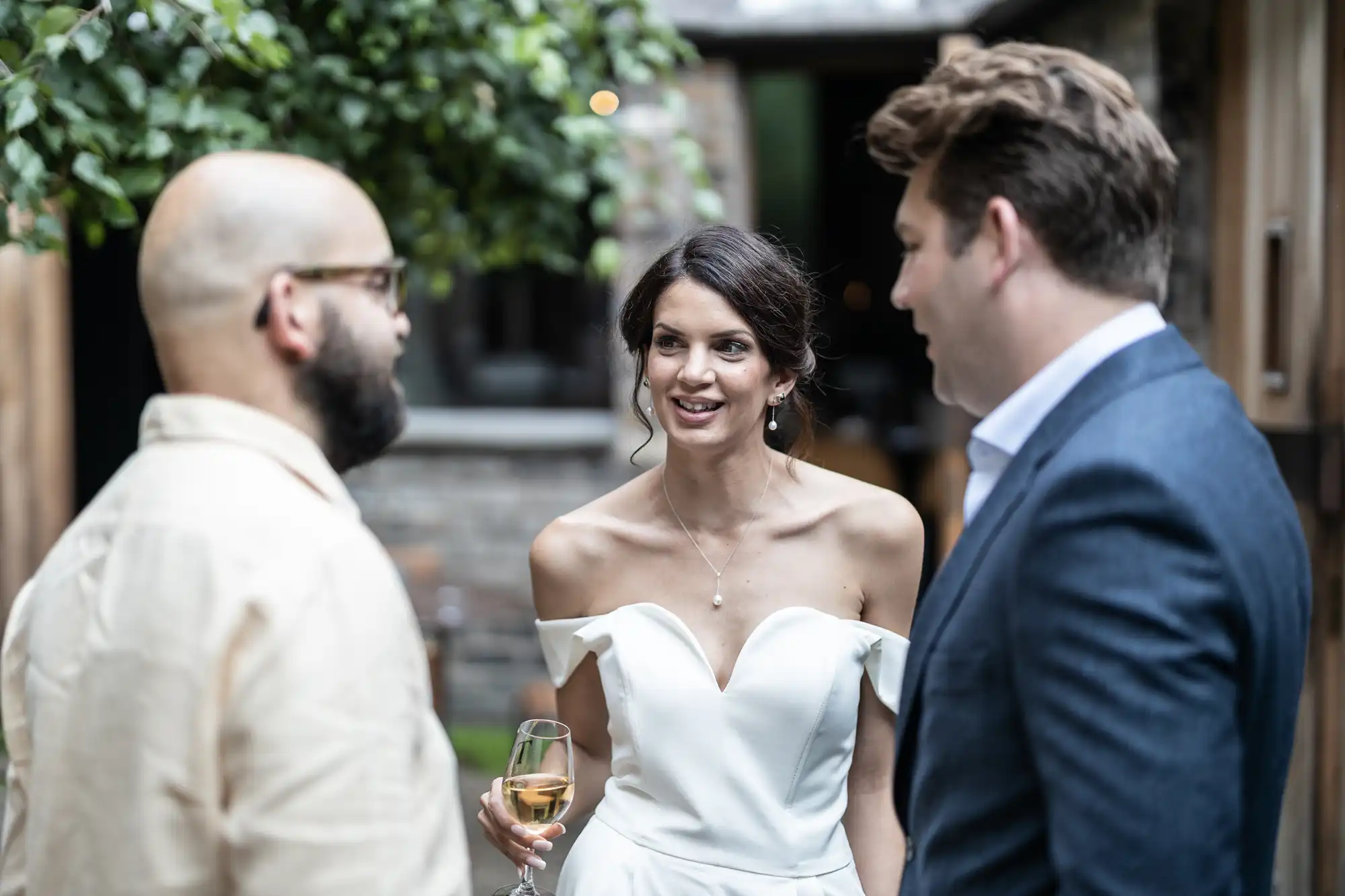 A bride in a white off-shoulder dress holding a glass of wine converses with two men in suits at an outdoor gathering.