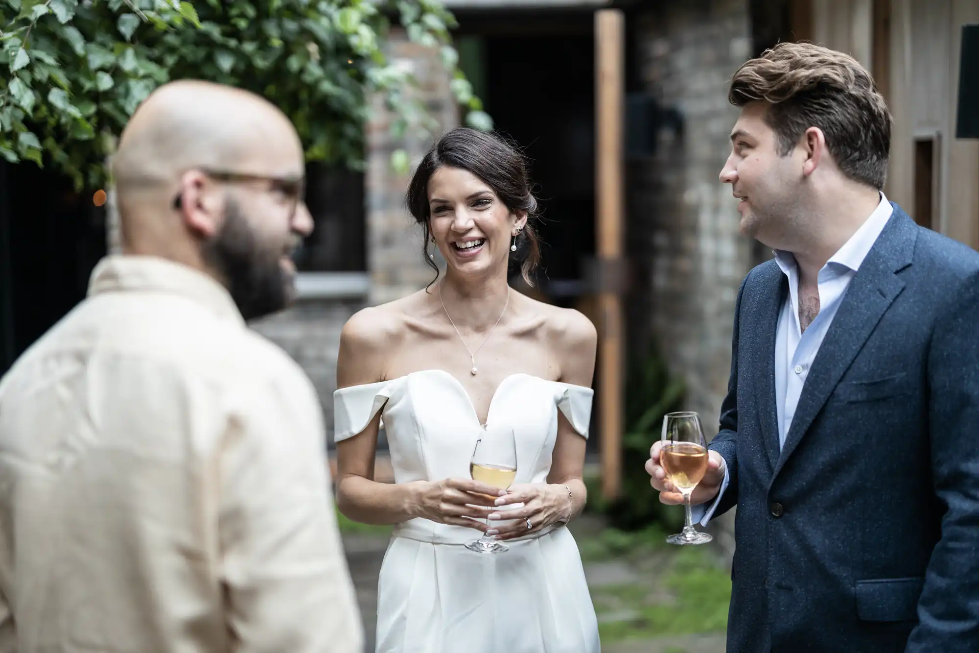 A bride in a white off-shoulder dress, holding a glass of wine, laughs joyfully while talking with two men in suits at an outdoor gathering.