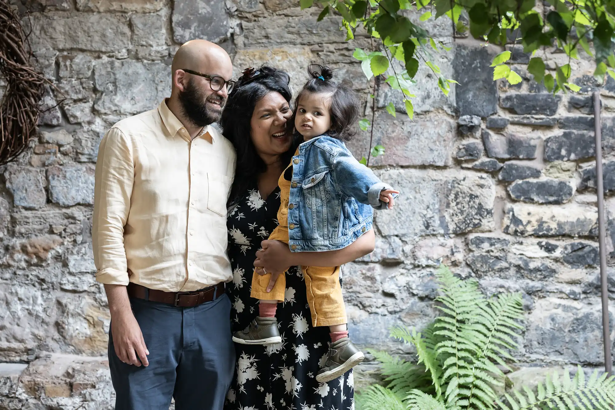 A family of three, including a bald man, a smiling woman, and a toddler, standing together against a stone wall with greenery.