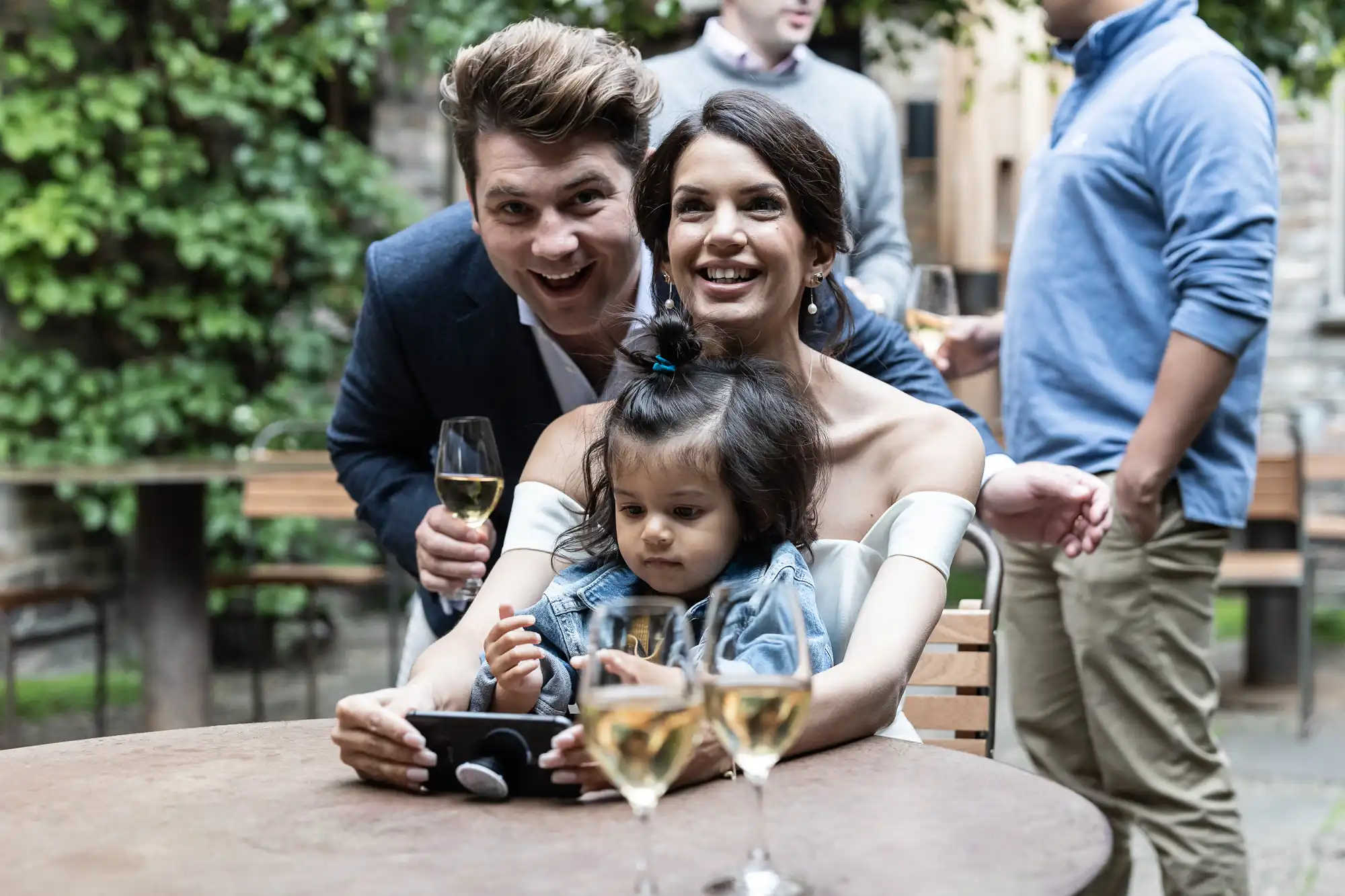 Happy couple with a young child sitting at a table at an outdoor gathering, holding glasses of wine and smiling.