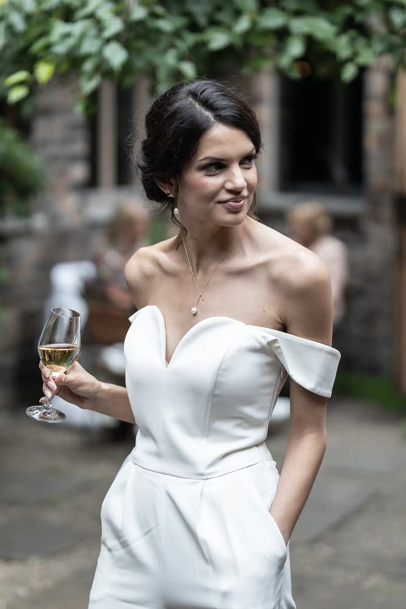 A woman in an elegant white off-the-shoulder dress holding a glass of wine at an outdoor event.