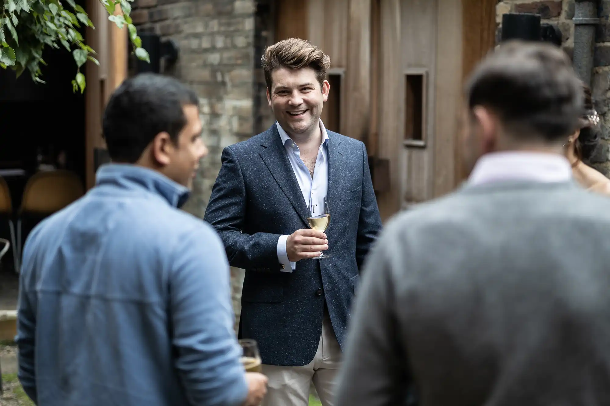 A smiling man in a blue blazer holding a wine glass while conversing with two other men at an outdoor gathering.