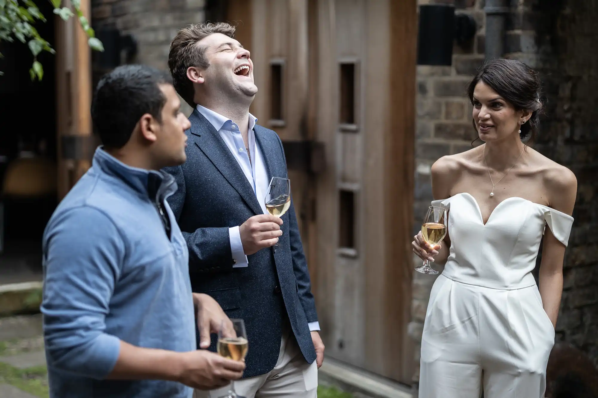 Three people at an outdoor event, laughing and holding glasses of drinks; one man in a casual shirt and jeans, another in a suit, and a woman in a white dress.