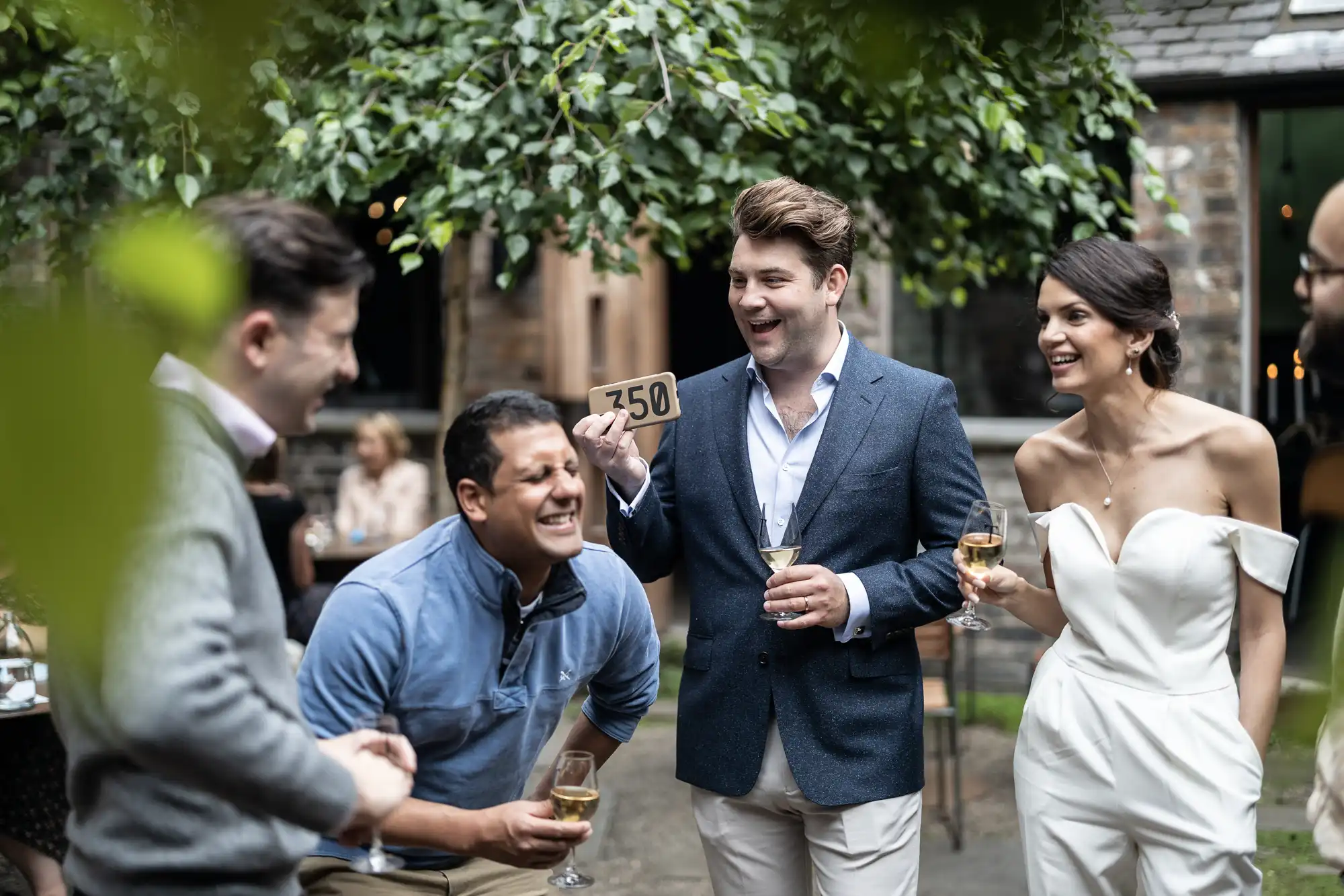 A group of people, including a bride and groom, enjoy a conversation and drinks at an outdoor wedding reception.