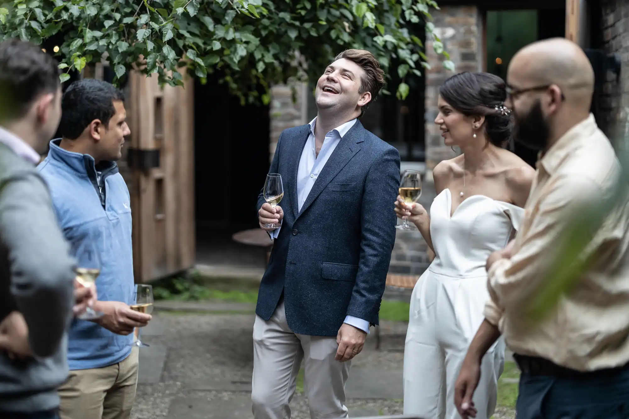 A joyful man in a blue blazer laughs while holding a wine glass, surrounded by a group of people in a courtyard, including a couple in wedding attire.