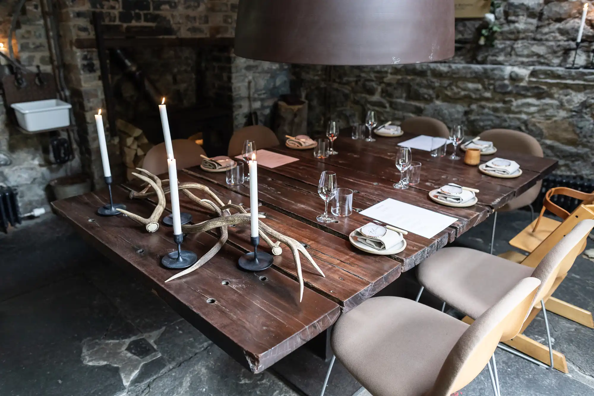A rustic dining table set for six, with candles, antlers, and simple dinnerware in a stone-walled room.