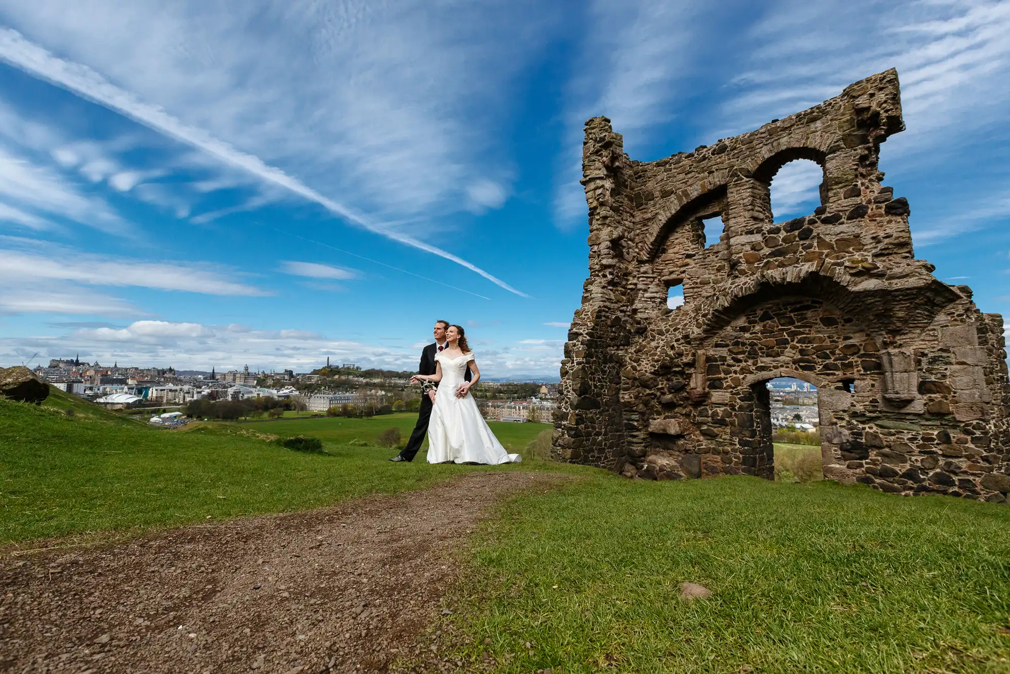 A bride and groom walking hand in hand near the ruins of an old stone structure, with a cityscape under a blue sky in the background.