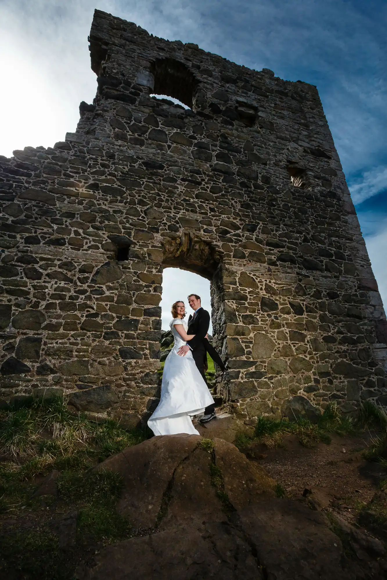 A bride and groom posing in the doorway of an ancient stone tower under a bright blue sky.