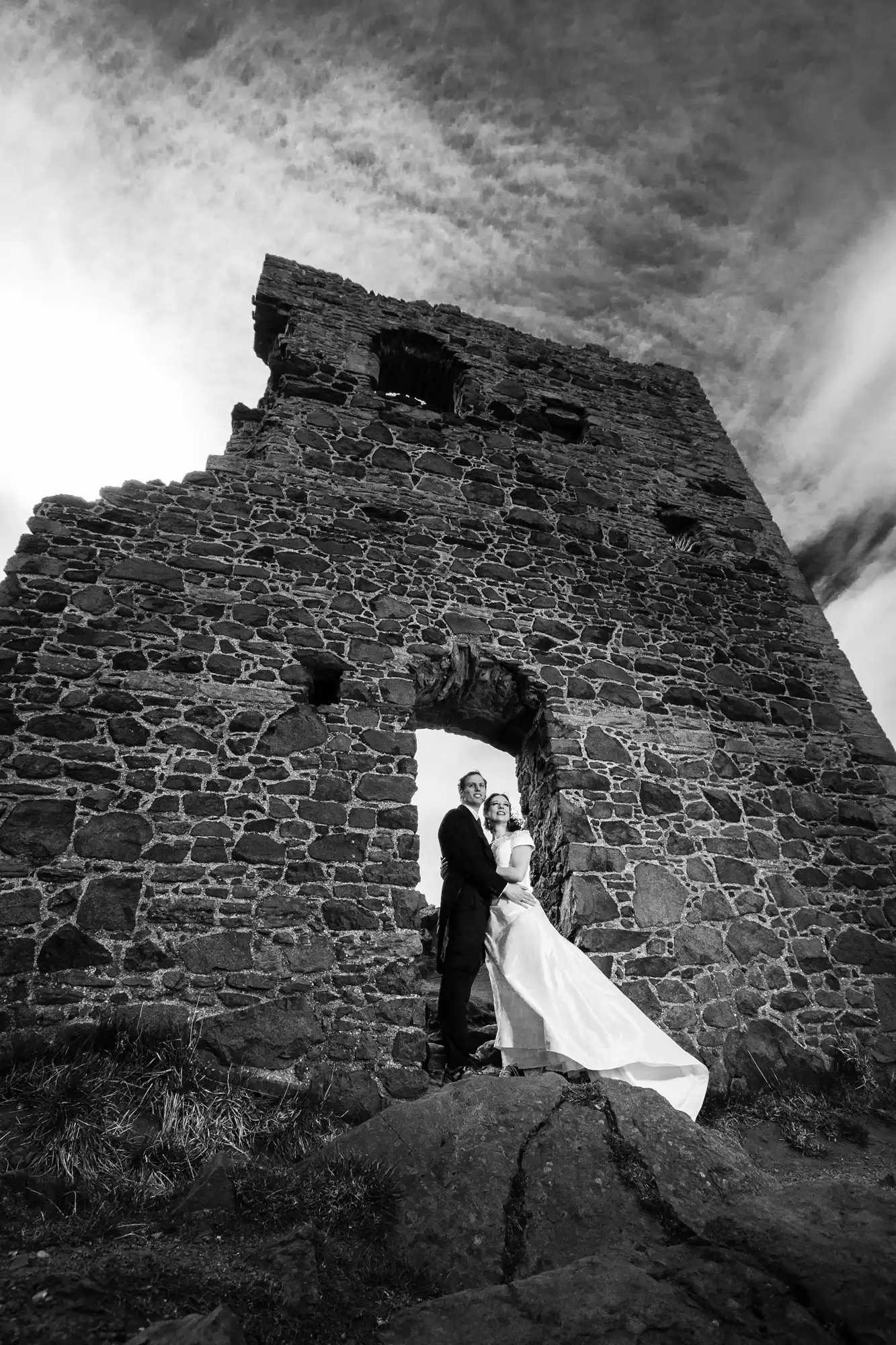 A bride and groom stand in an embrace under the archway of a ruined stone tower, with a dramatic sky overhead.