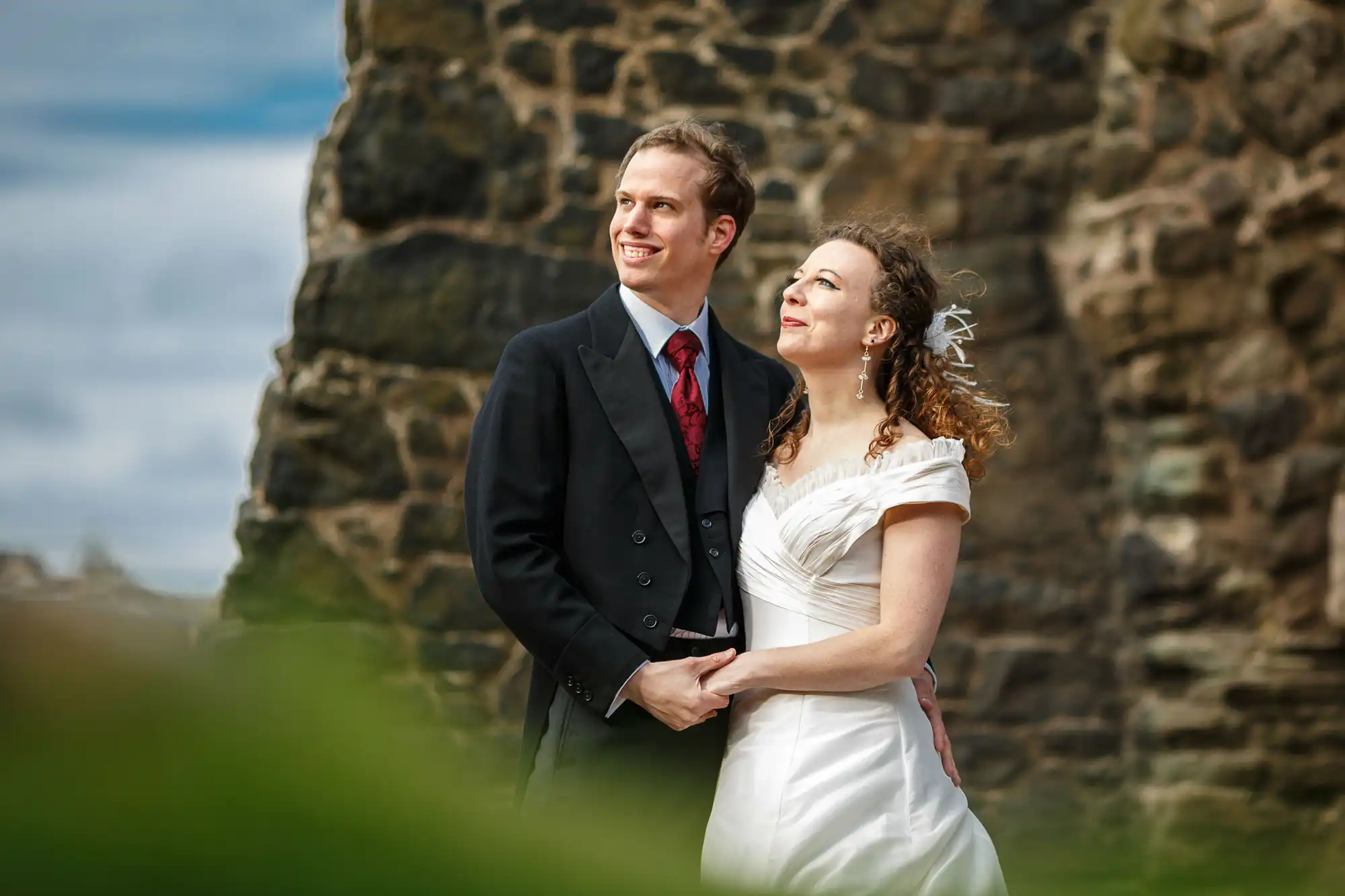 A bride and groom holding hands and smiling, standing outdoors with an old stone structure in the background and greenery in the foreground.