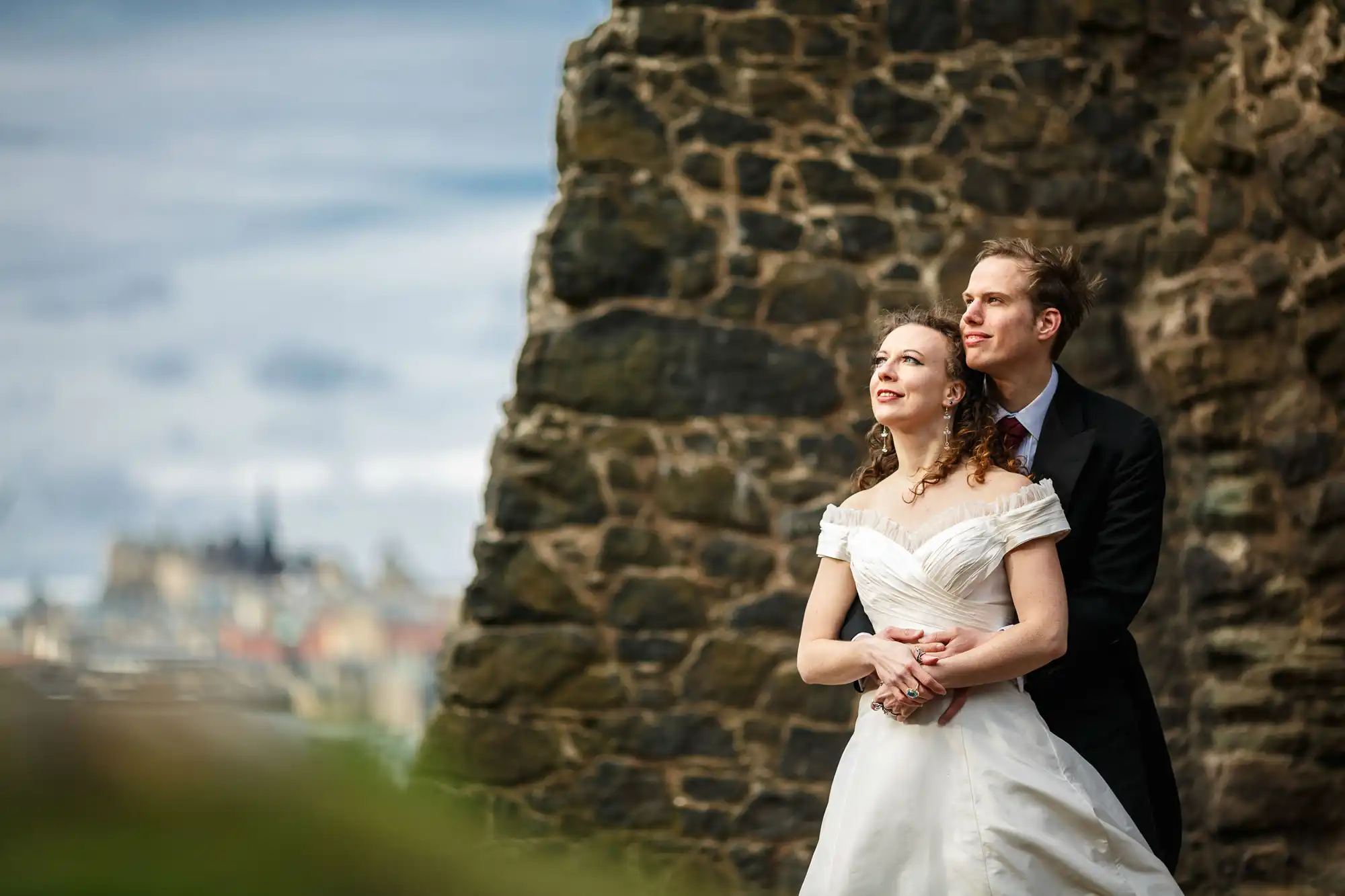 A bride and groom stand together near old stone ruins, looking into the distance with a cityscape in the background.