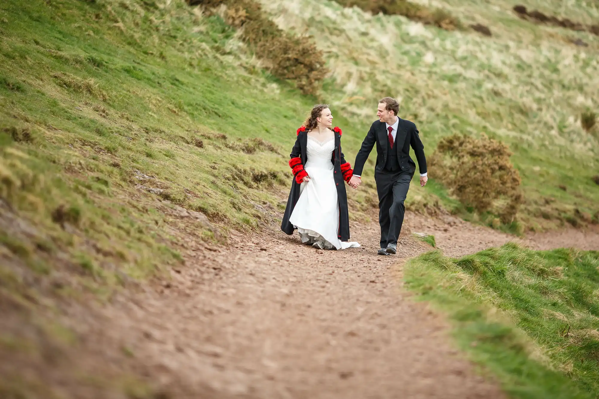 A bride in a white dress and a groom in a black suit walking together on a rural path, with grassy hills around them.
