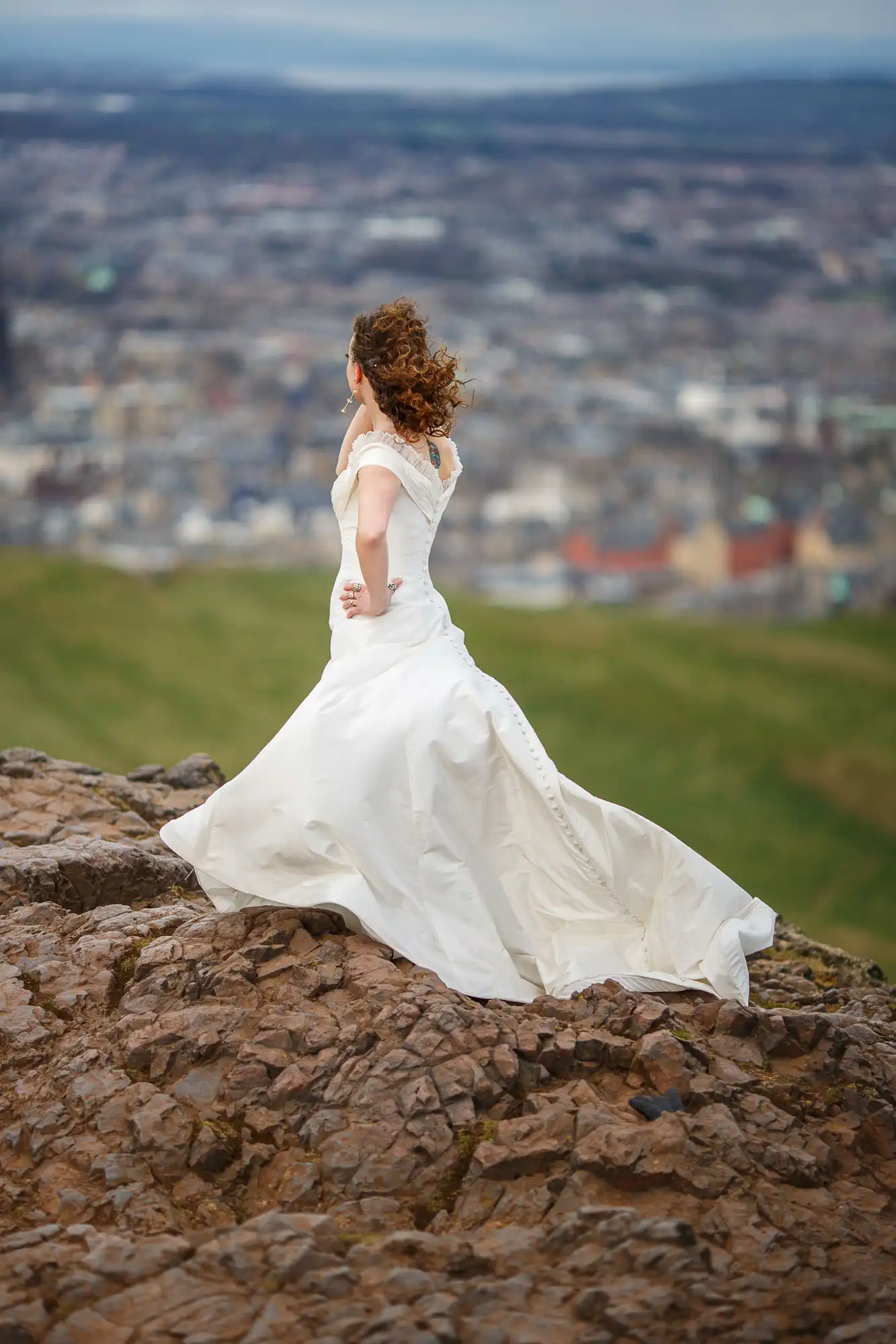 A woman in a white wedding dress stands on a rocky hilltop overlooking a cityscape, her curly hair blowing in the wind.