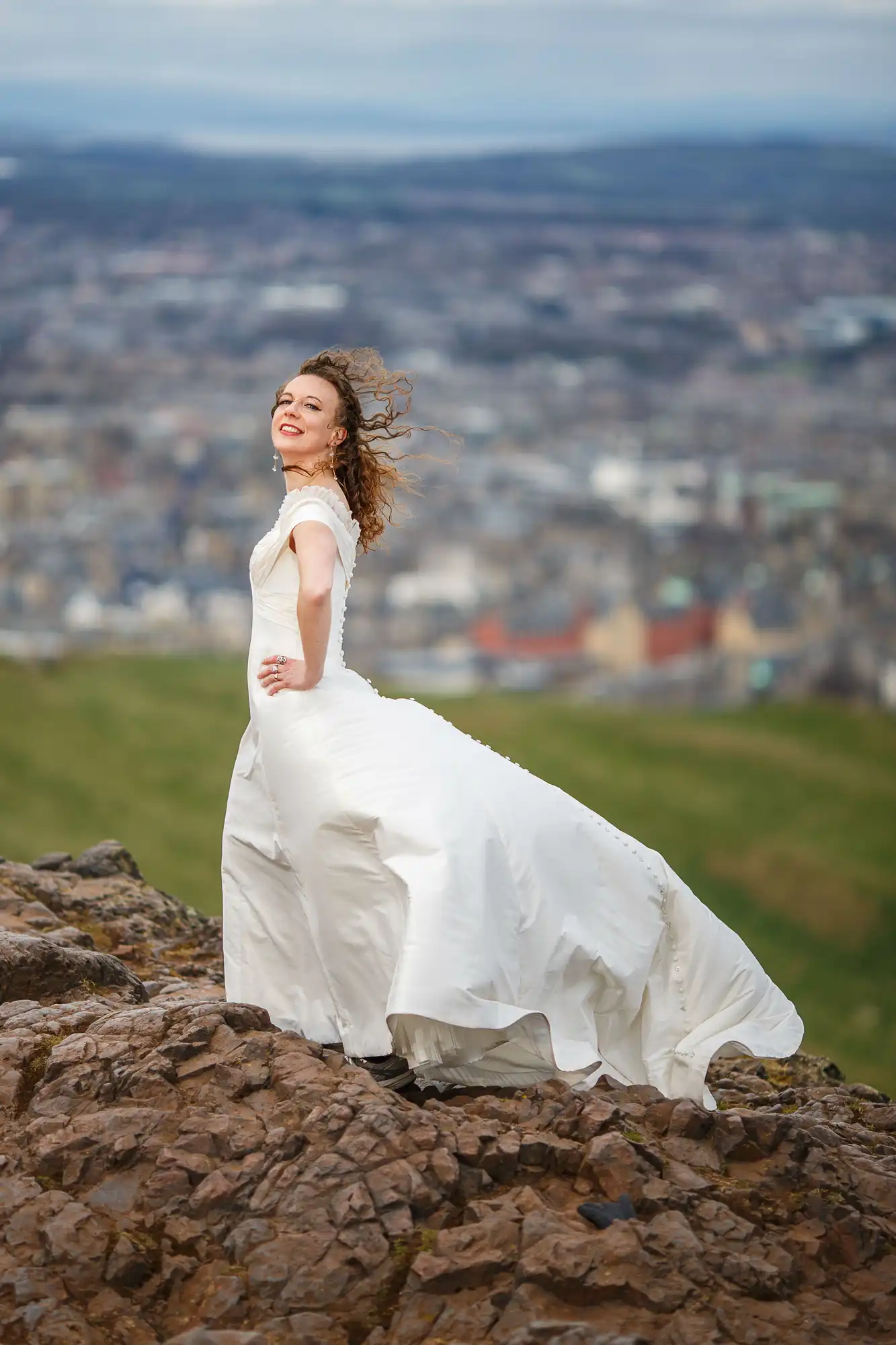 A joyful bride in a white dress stands on a rocky hilltop, her hair blowing in the wind, with a panoramic view of a cityscape in the background.
