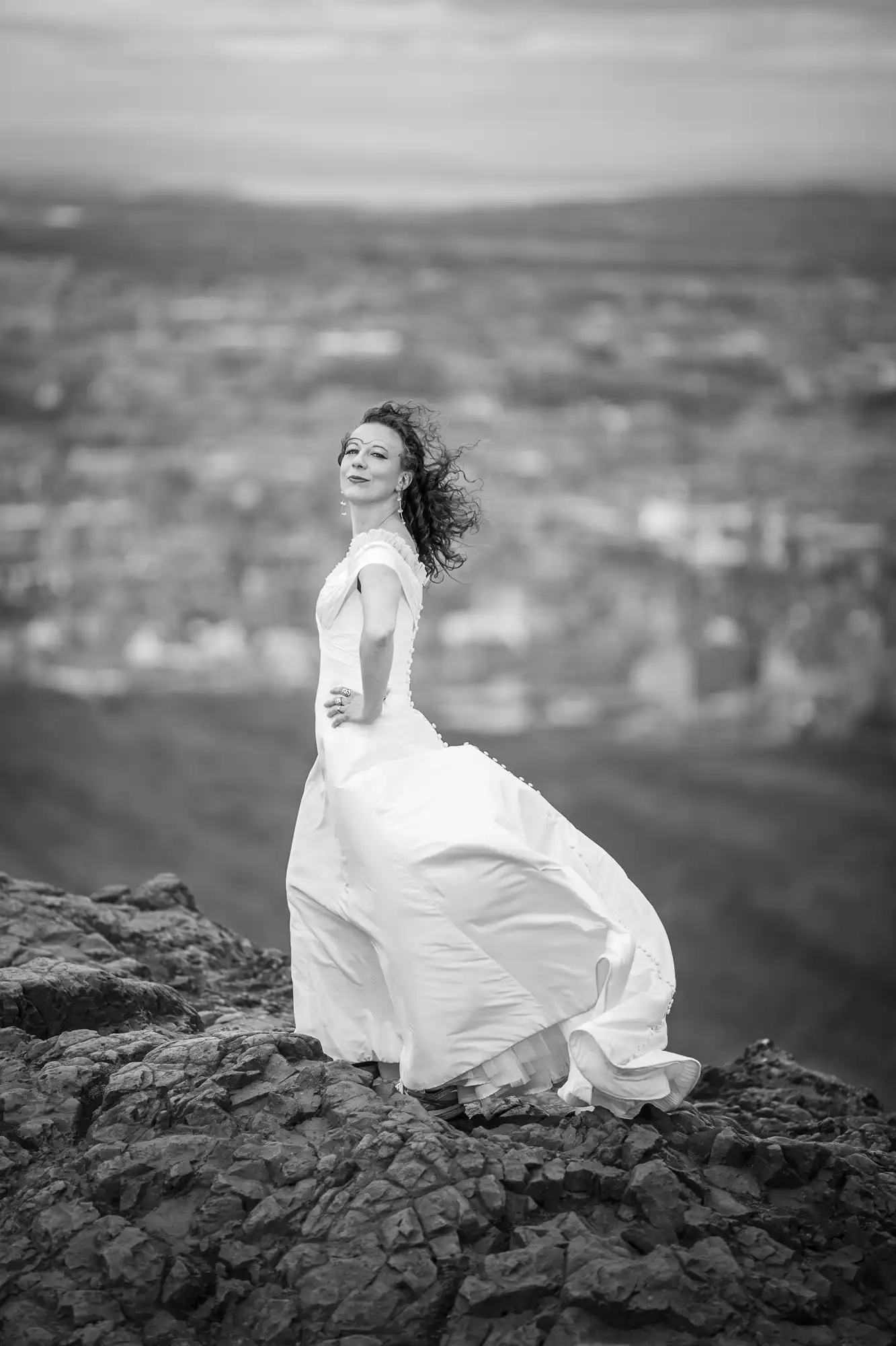 A woman in a white dress stands on a rocky peak, her hair blowing in the wind, with a blurred cityscape in the background, captured in black and white.