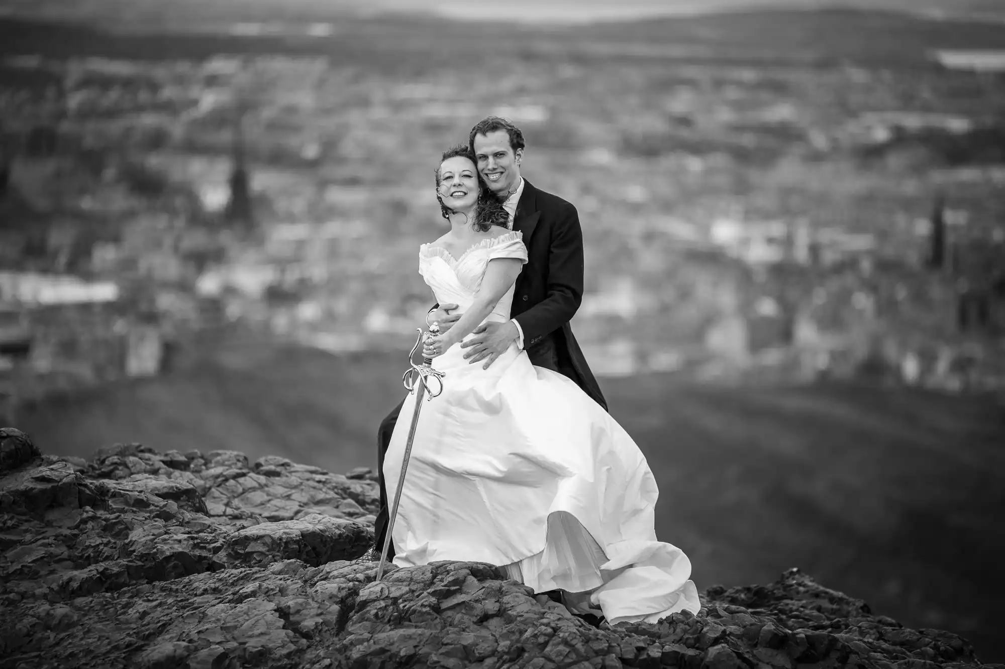 A black and white image of a bride and groom smiling and standing on rocky terrain with a blurred cityscape in the background.