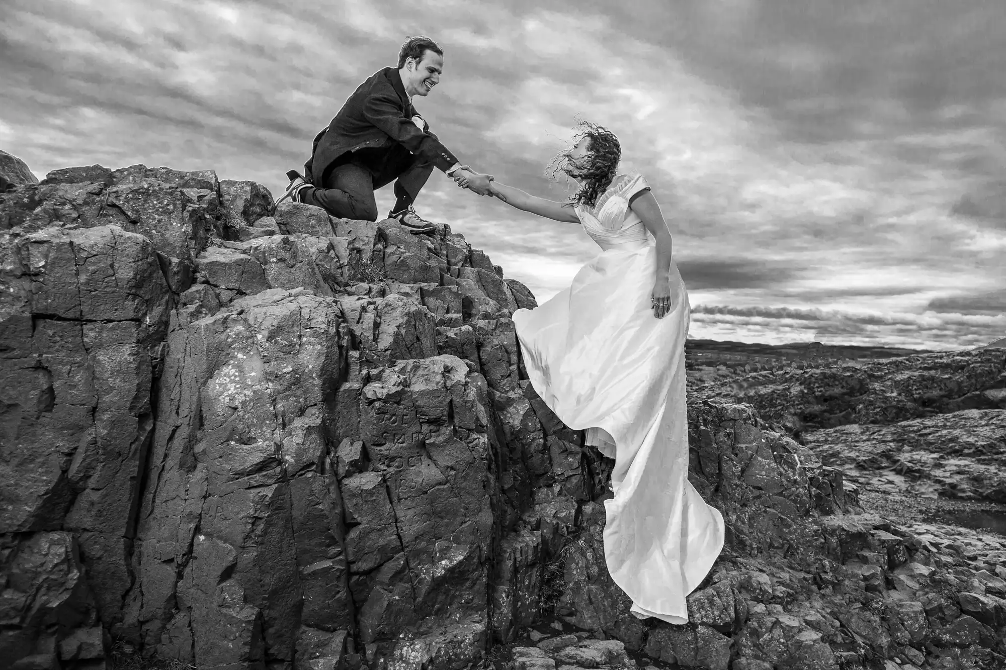A bride in a white dress and a groom in a suit hold hands on opposite rocky cliffs under a cloudy sky.