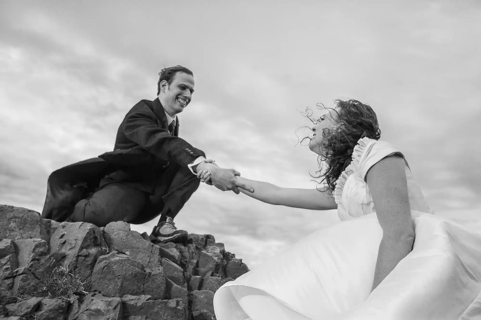 A black and white photo of a joyful bride and groom holding hands, with the groom on a higher rock ledge, pulling the bride upwards as she climbs.