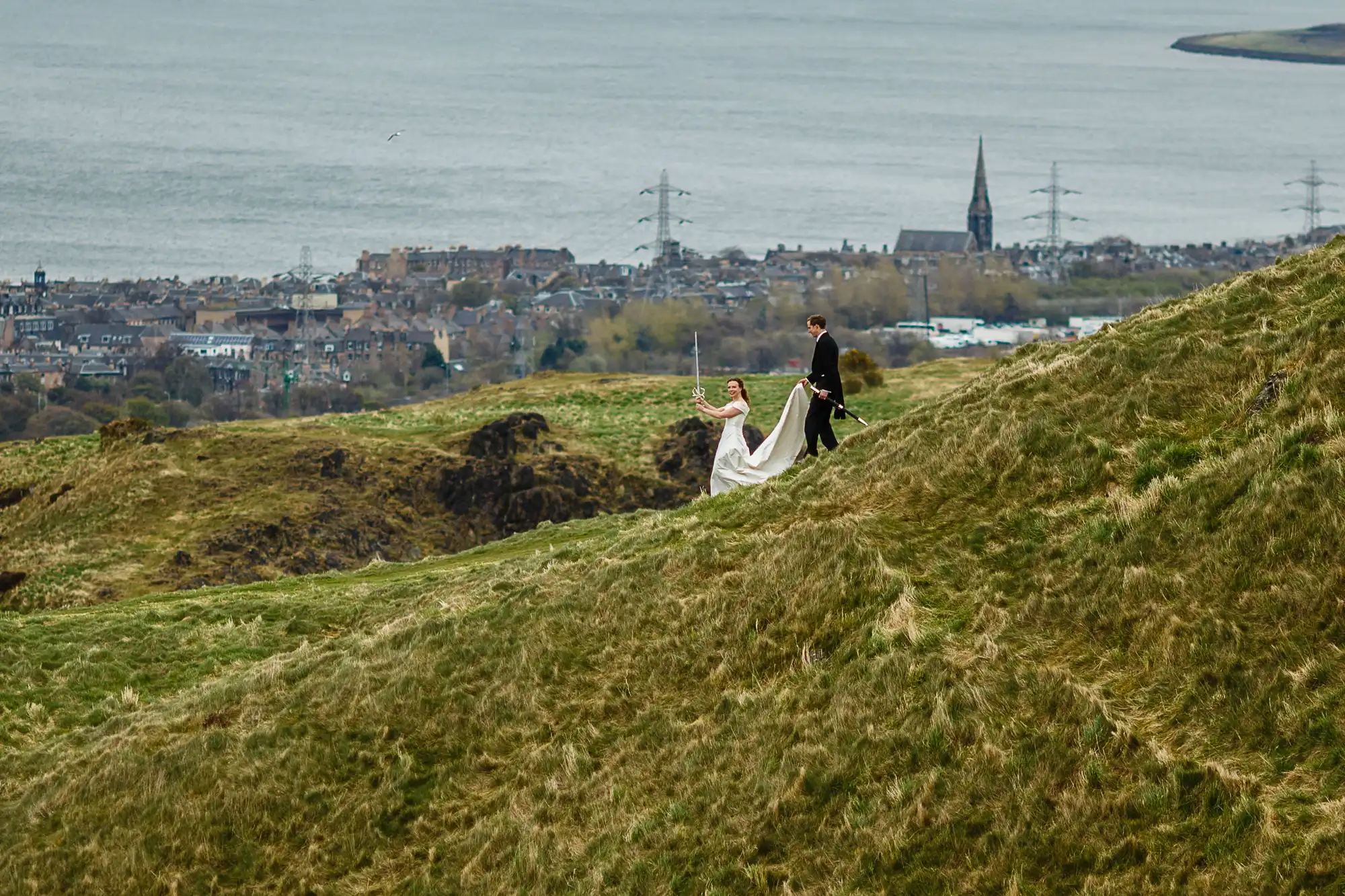 A couple in wedding attire walk hand in hand on a grassy hill, with a coastal town and water in the background.