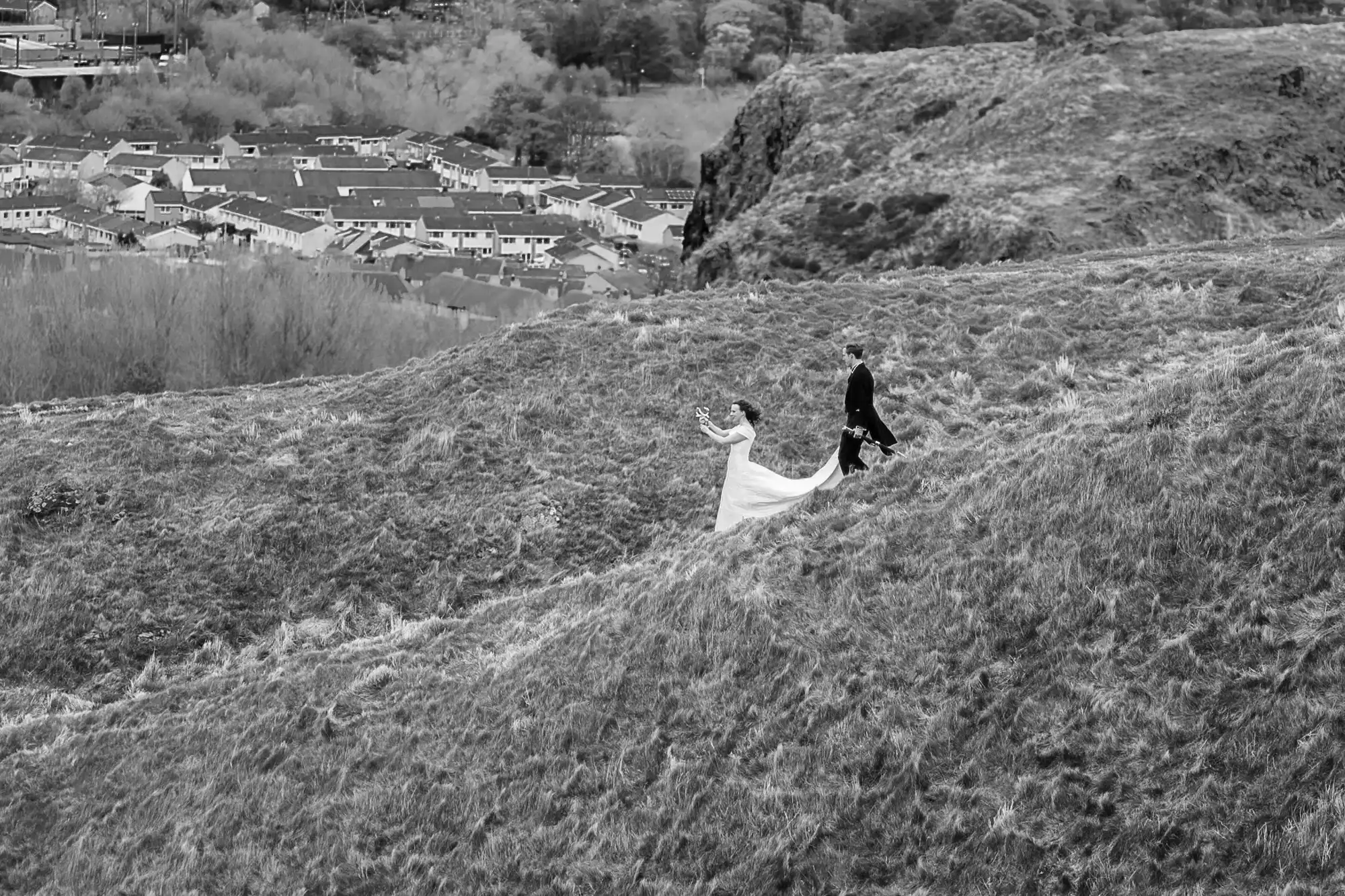 A bride and groom walking up a grassy hill, with the bride's long white dress trailing behind, against a backdrop of a small town.