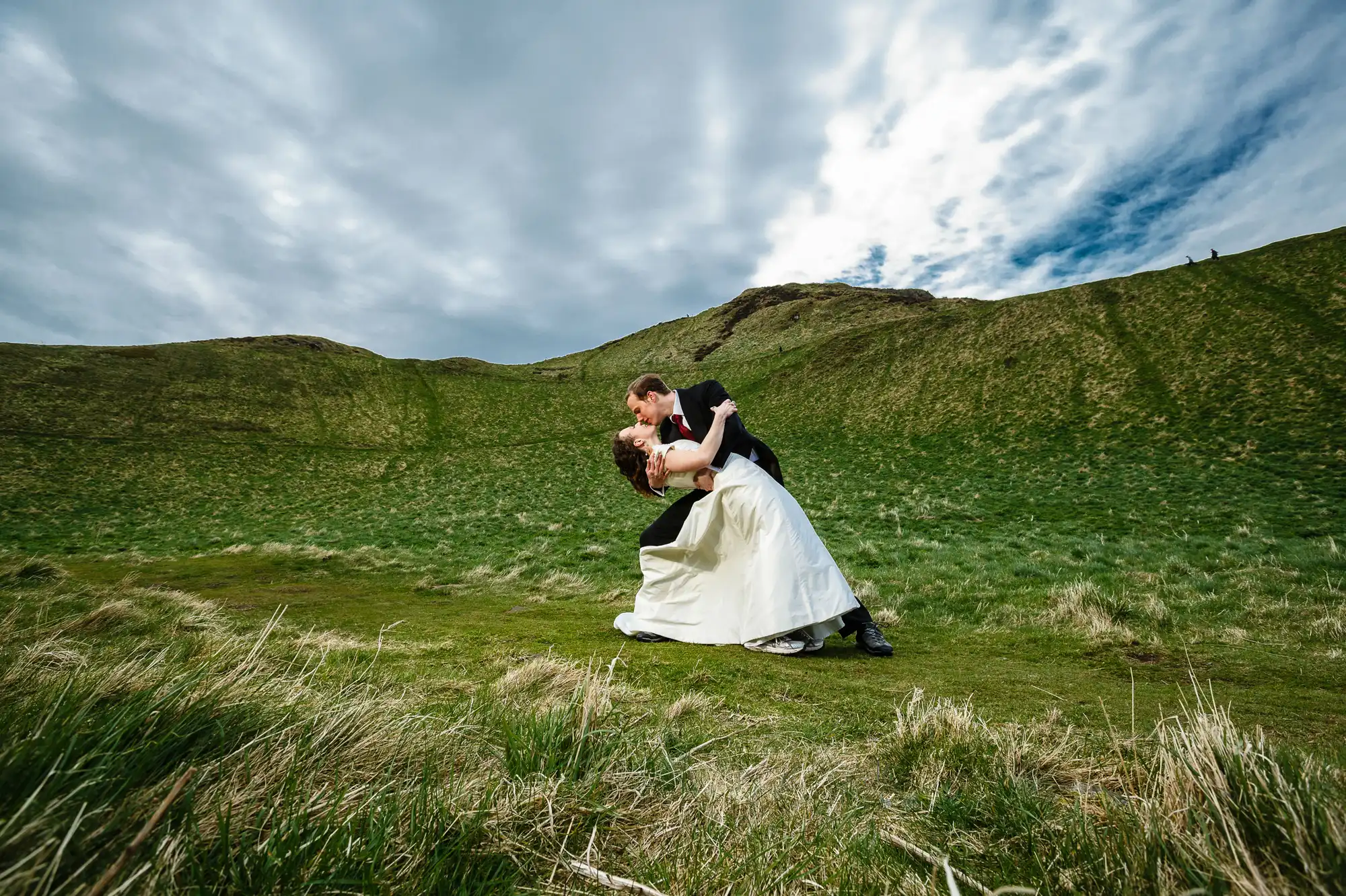 A couple in wedding attire dancing in a lush green field under a dramatic cloudy sky.