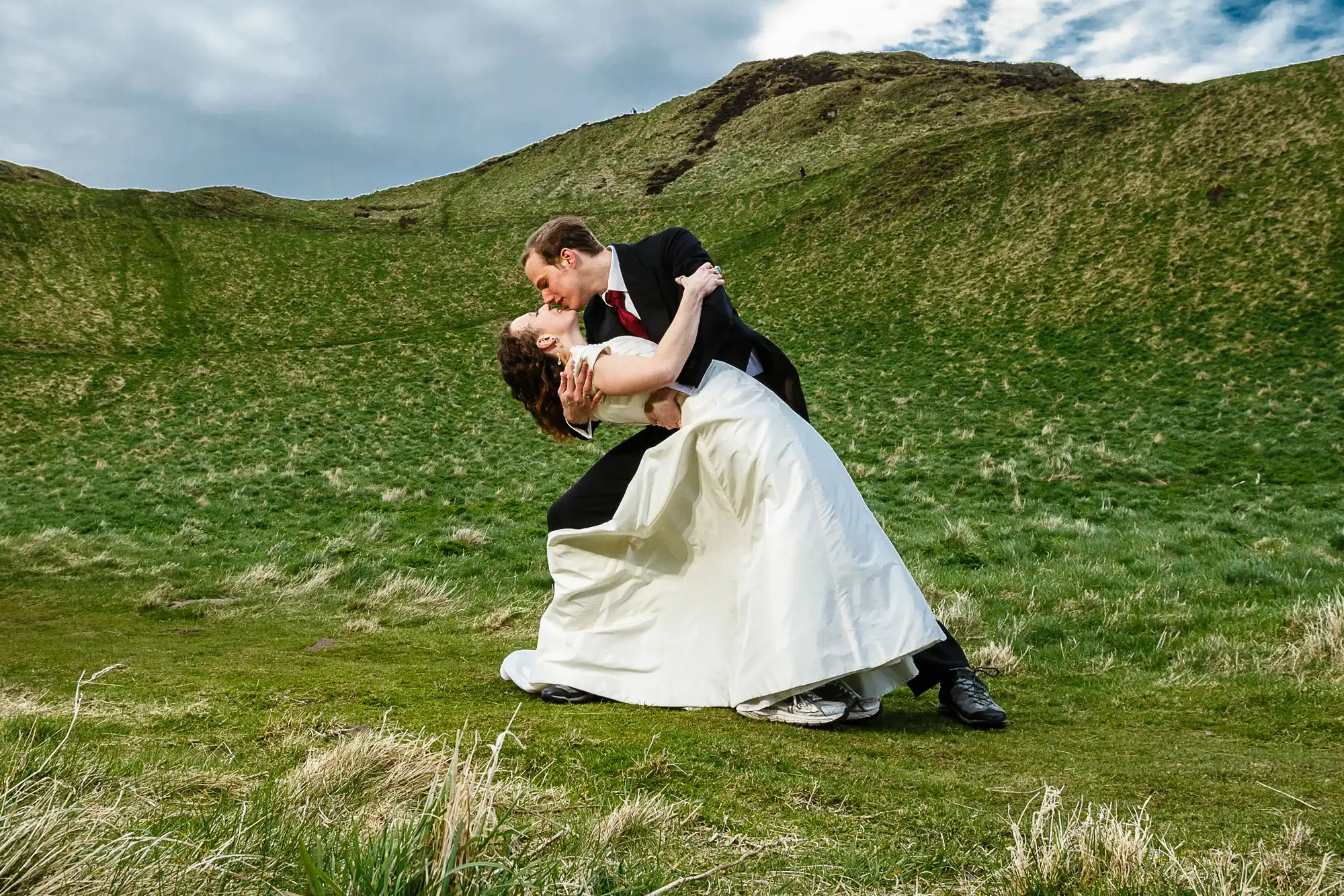 A bride and groom sharing a kiss while performing a dip in a lush, green meadow with rolling hills in the background.