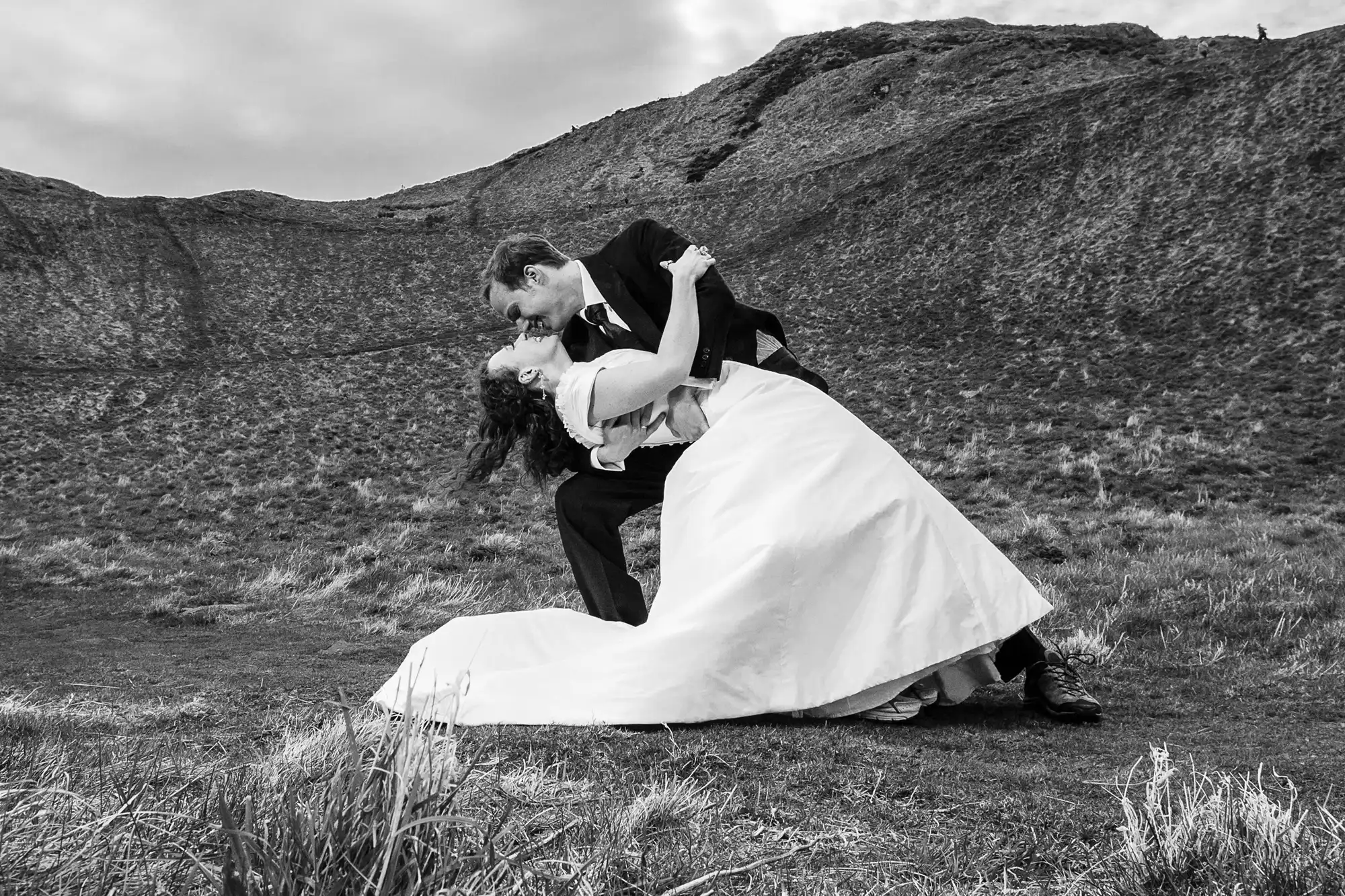A bride and groom sharing a dramatic kiss outdoors with a rugged hill in the background, all in black and white.